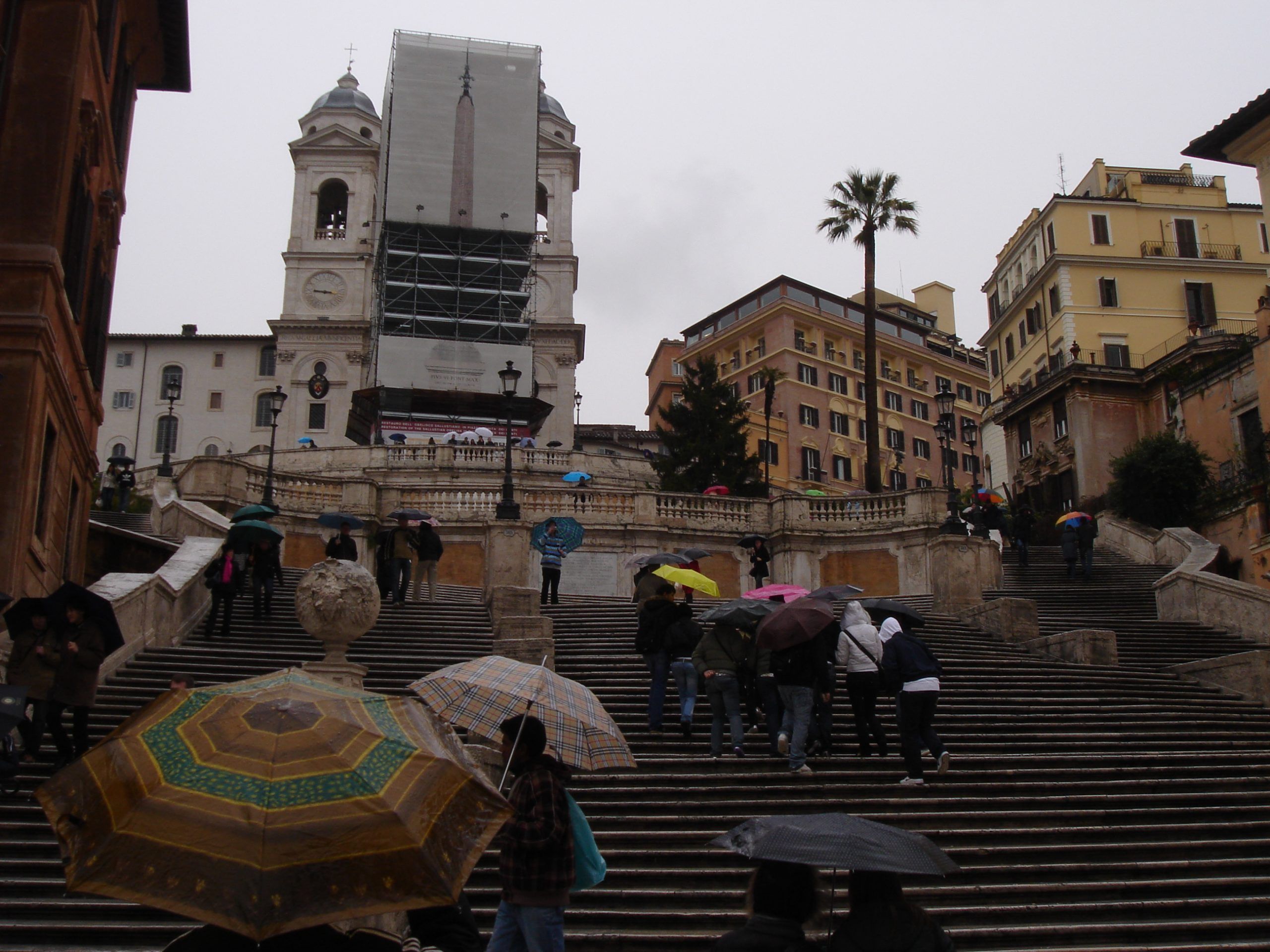 Tourists with umbrellas ascending the Spanish Steps in Rome on a rainy day, with the Trinità dei Monti church under scaffolding at the top.