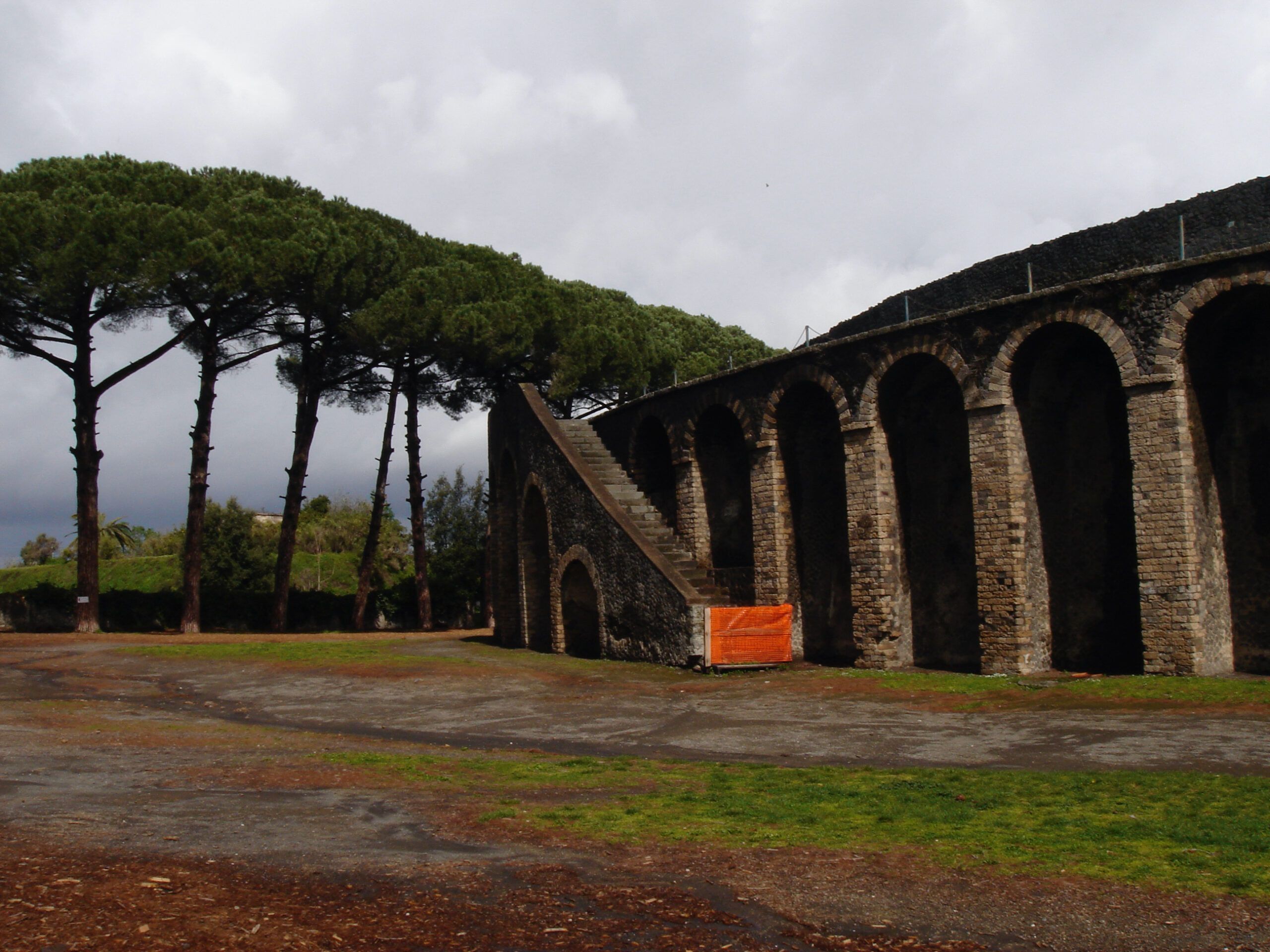 Outer wall of Pompeii’s amphitheatre with dramatic rows of arches and pine trees, under a brightening sky after rain.