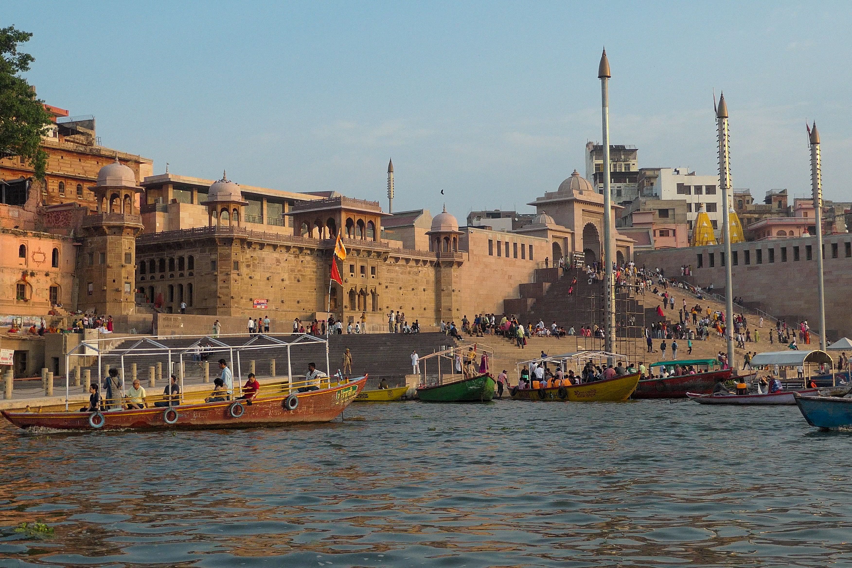 Boats and people in front of the historic sandstone buildings and steps of a Varanasi ghat in the morning light.