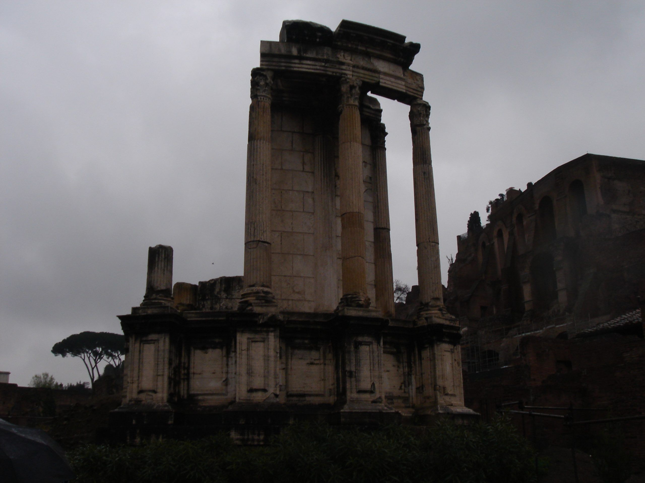 The three remaining columns of the Temple of Vesta in the Roman Forum, silhouetted dramatically against a grey, overcast sky.