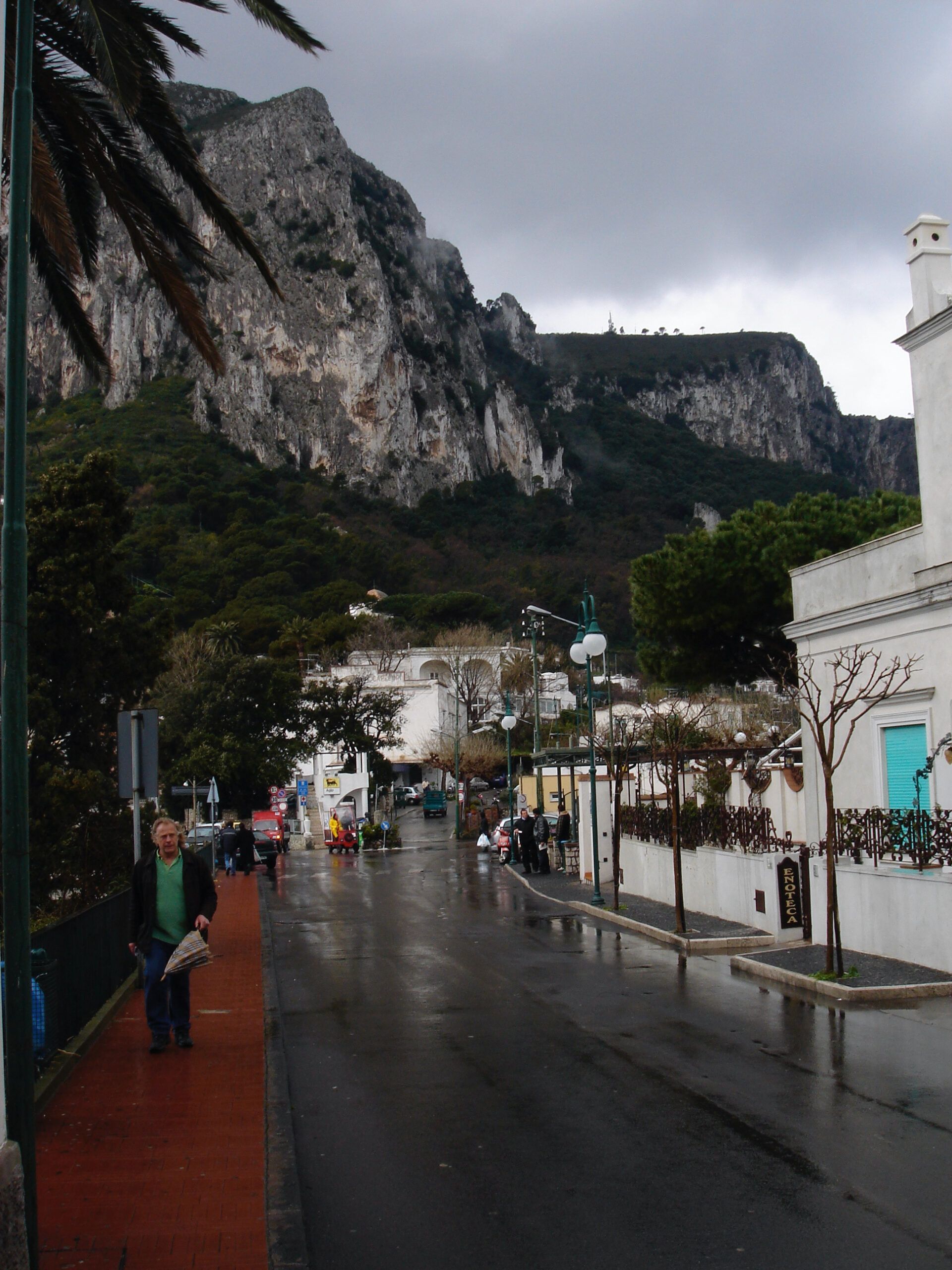 A street in Capri on a rainy day, leading toward steep limestone cliffs with buildings and trees lining the road.