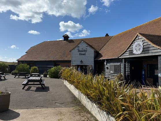 A pub with dark timber and whitewashed walls under a reddish-brown tiled roof, labeled “The Beachy Head.” Picnic tables sit on the paved courtyard in front, under a bright blue sky with scattered clouds.