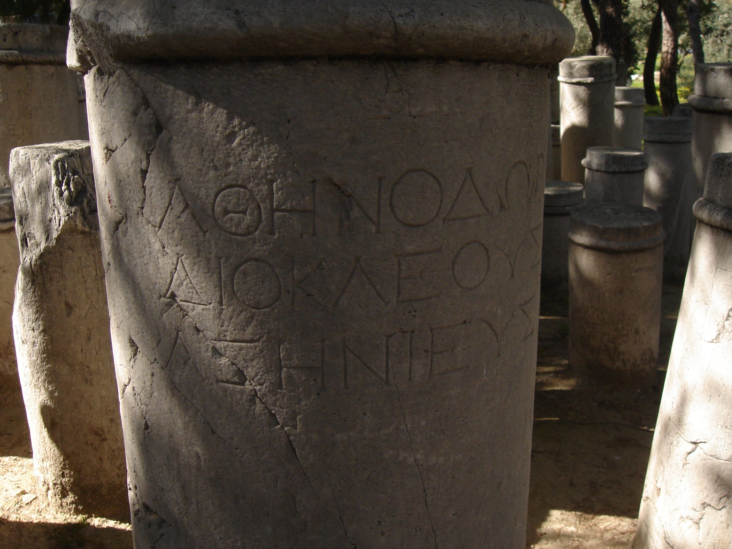 A cylindrical stone grave marker inscribed in Ancient Greek, surrounded by other similar stones in dappled light.