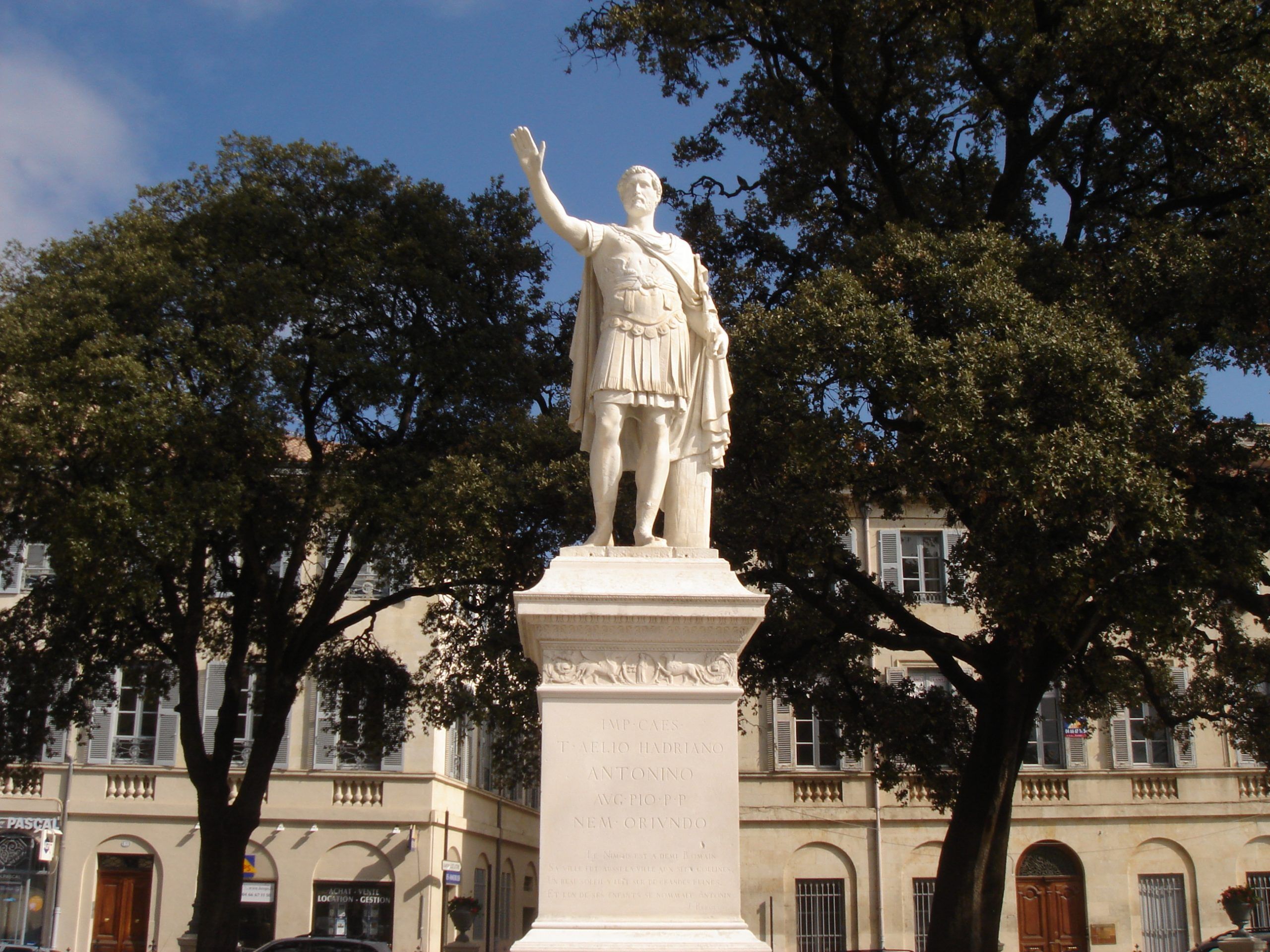 Statue of Antoninus Pius standing on a plinth in a square in Nîmes, surrounded by trees and buildings.