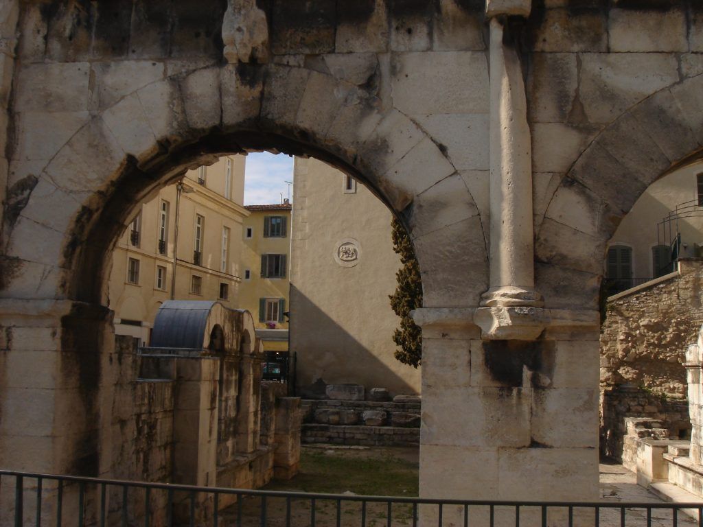 Roman stone archway framing a view of modern buildings in Nîmes, with archaeological remains in the foreground.