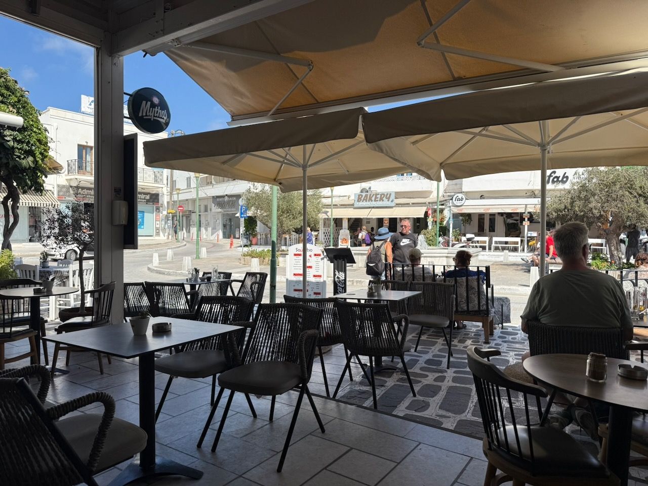 The outdoor seating area of a café, looking out onto a small square surounded by shops and restaurants. It's very sunny, and there are a few people sitting at the tables.