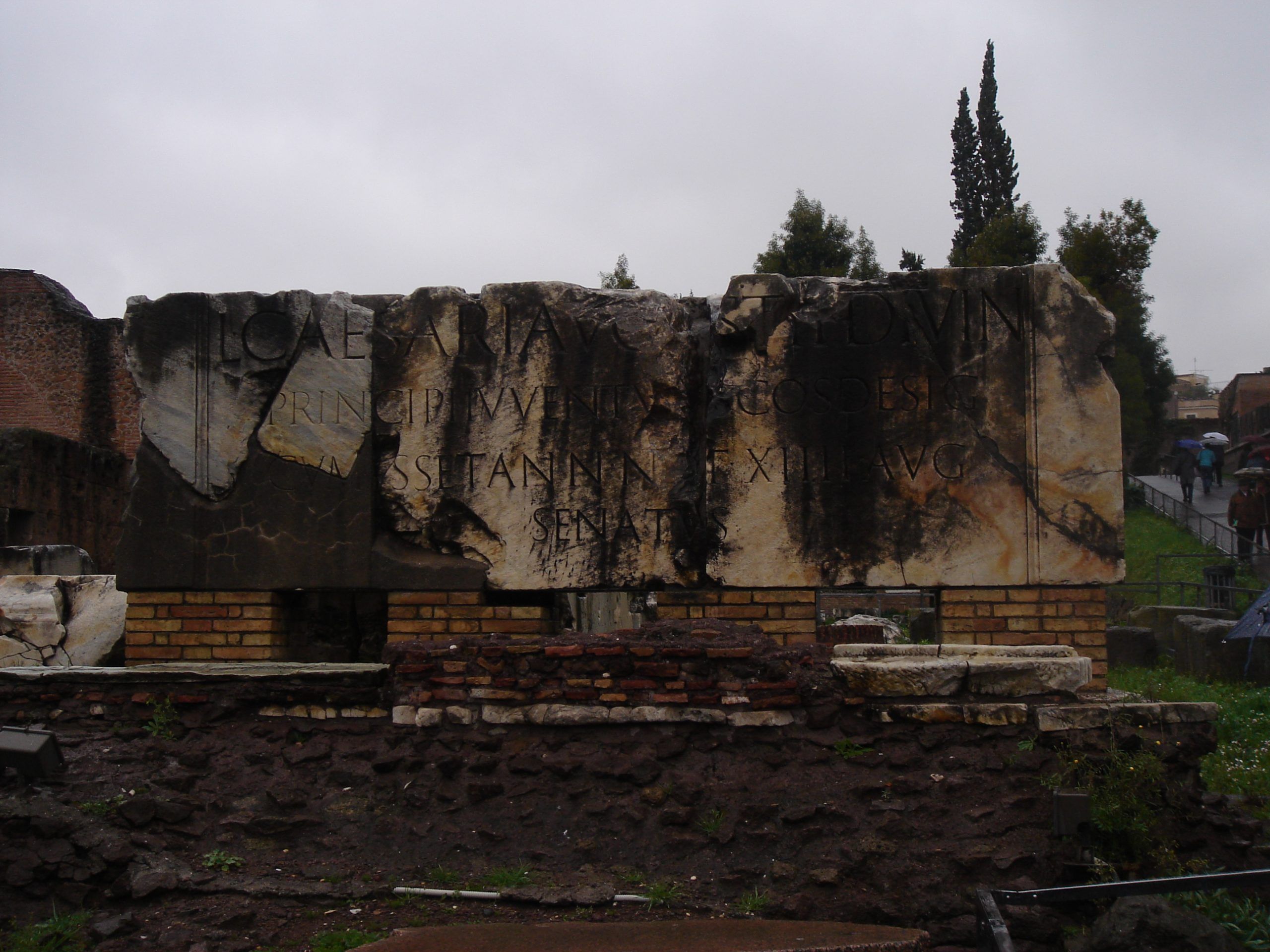 Fragmented marble inscription slab at the Roman Forum, scorched and mounted on a brick support, with traces of Latin text honoring an emperor.