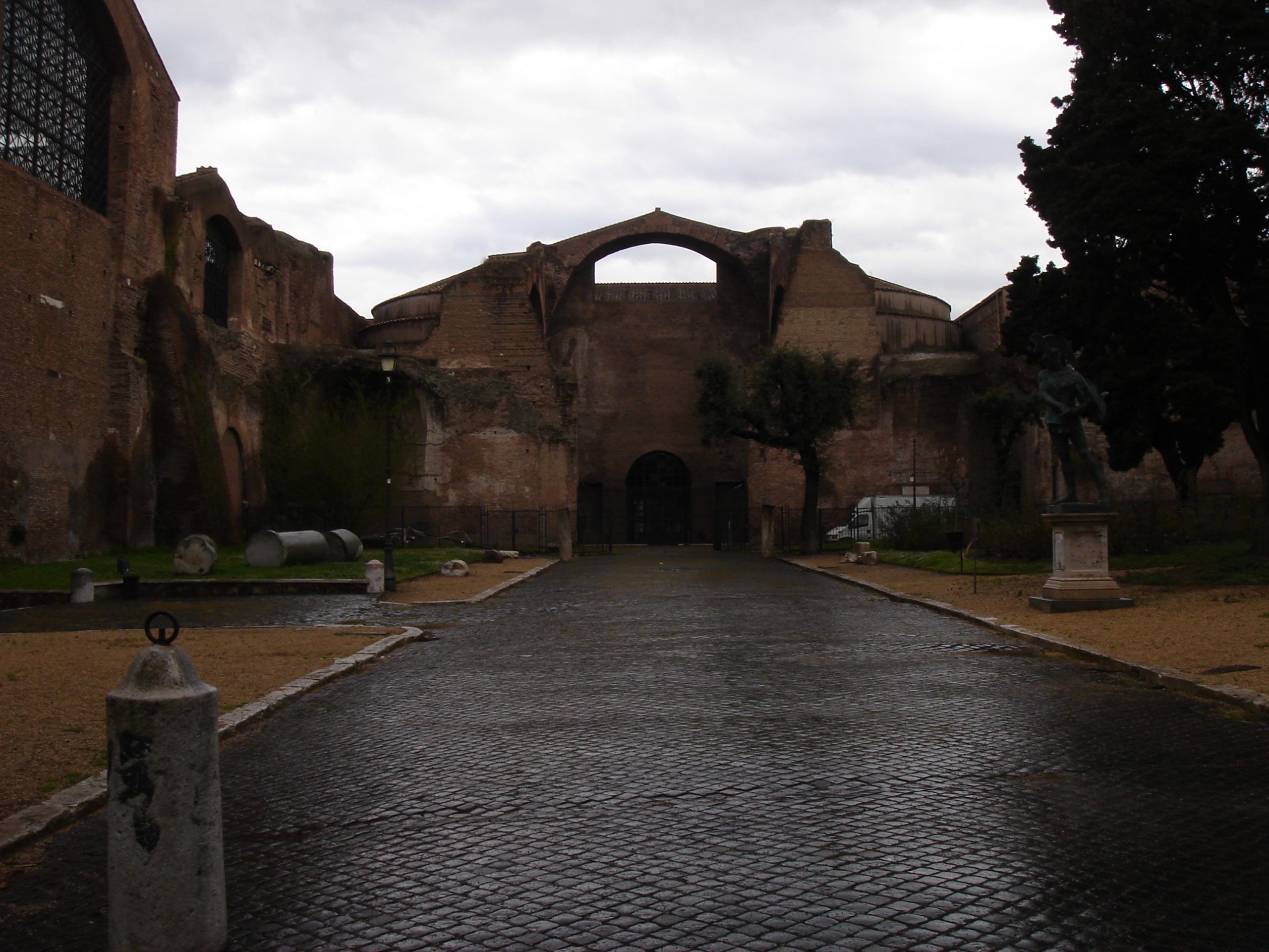 View of the Baths of Diocletian in Rome on a wet day, with scattered ruins, trees, and a statue near the path leading to the entrance.