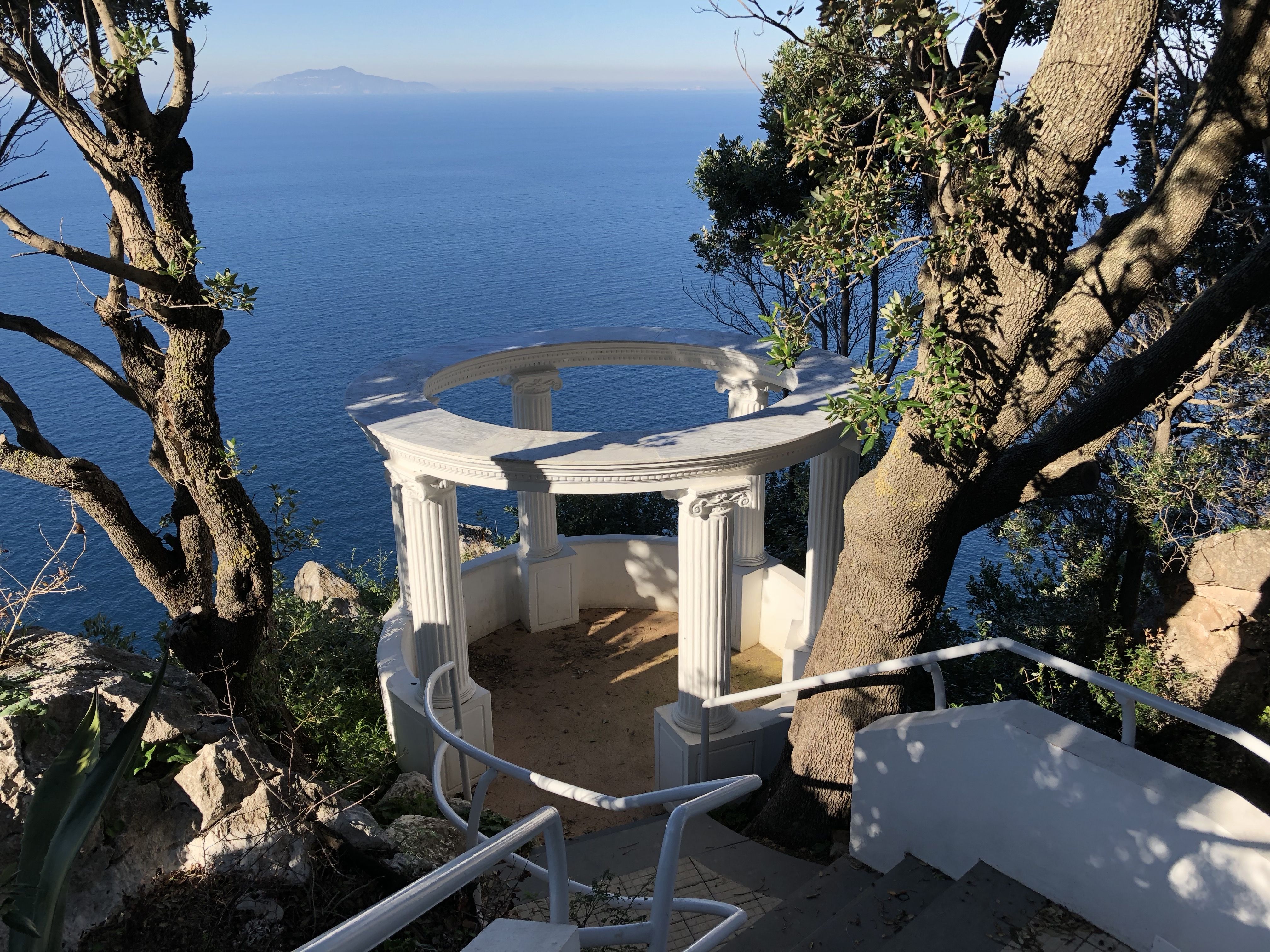 Marble-columned belvedere perched on the cliffs of Capri, seen from above through surrounding trees, with the sea stretching into the distance.