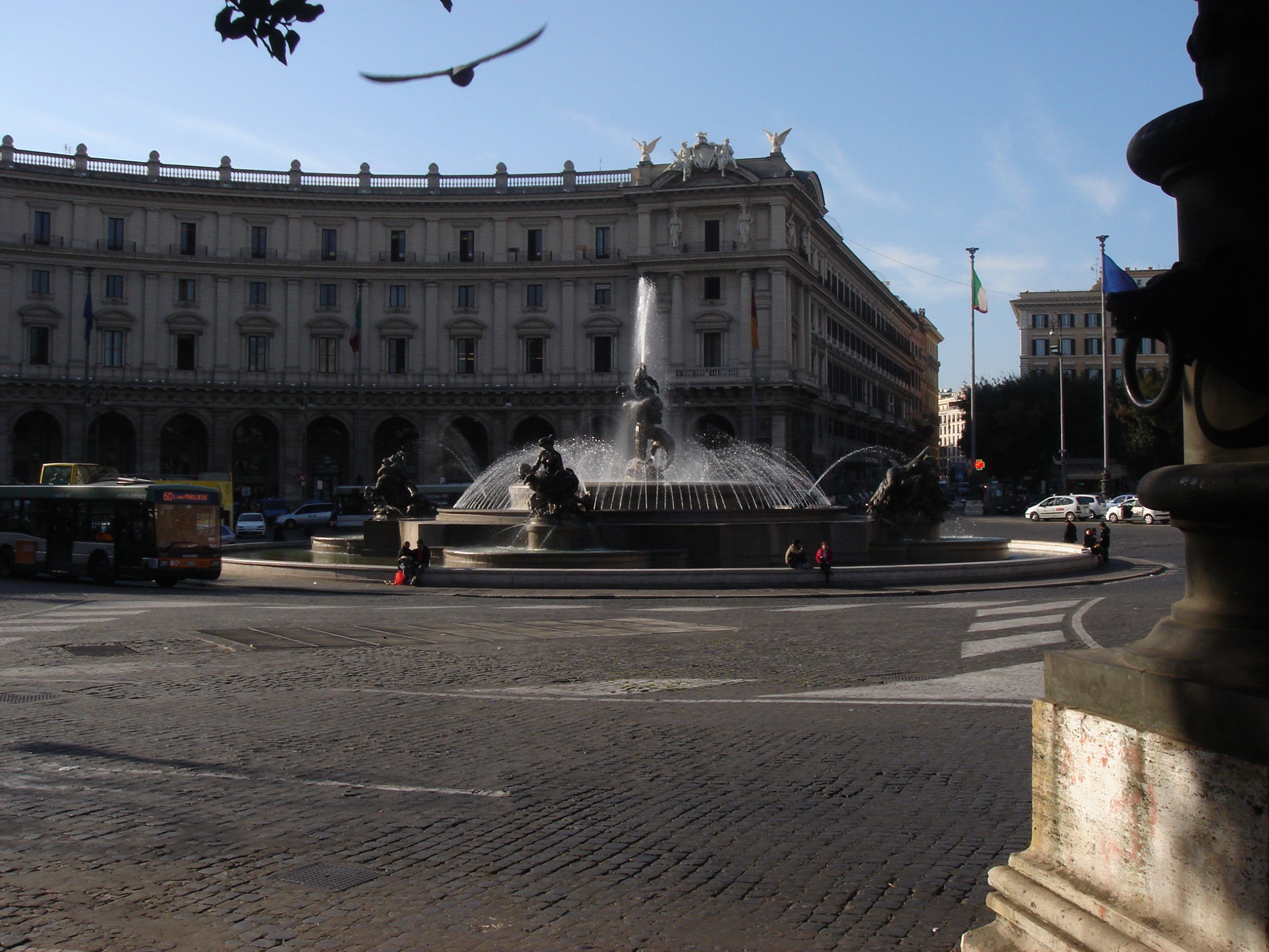 A wide view of the Fountain of the Naiads in Piazza della Repubblica, Rome, with neoclassical buildings and buses in the background.