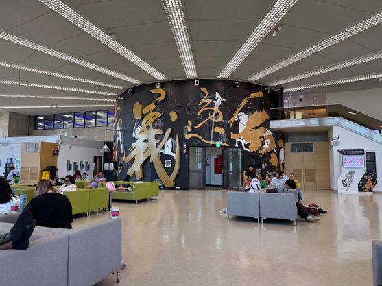 A huge room full of sleepy travellers on sofas. The ceiling is high and on the wall is an ornate decoration in gold, white and black, featuring horses and riders and massive gold Chinese characters