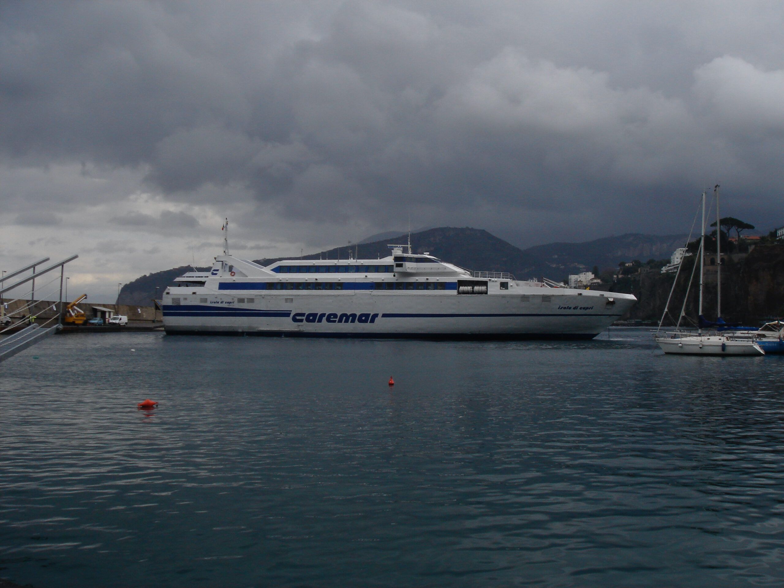 A Caremar ferry named 'Isola di Capri' docked at Porto Grande under a cloudy sky, with sailboats and a mountainous coastline in the background.