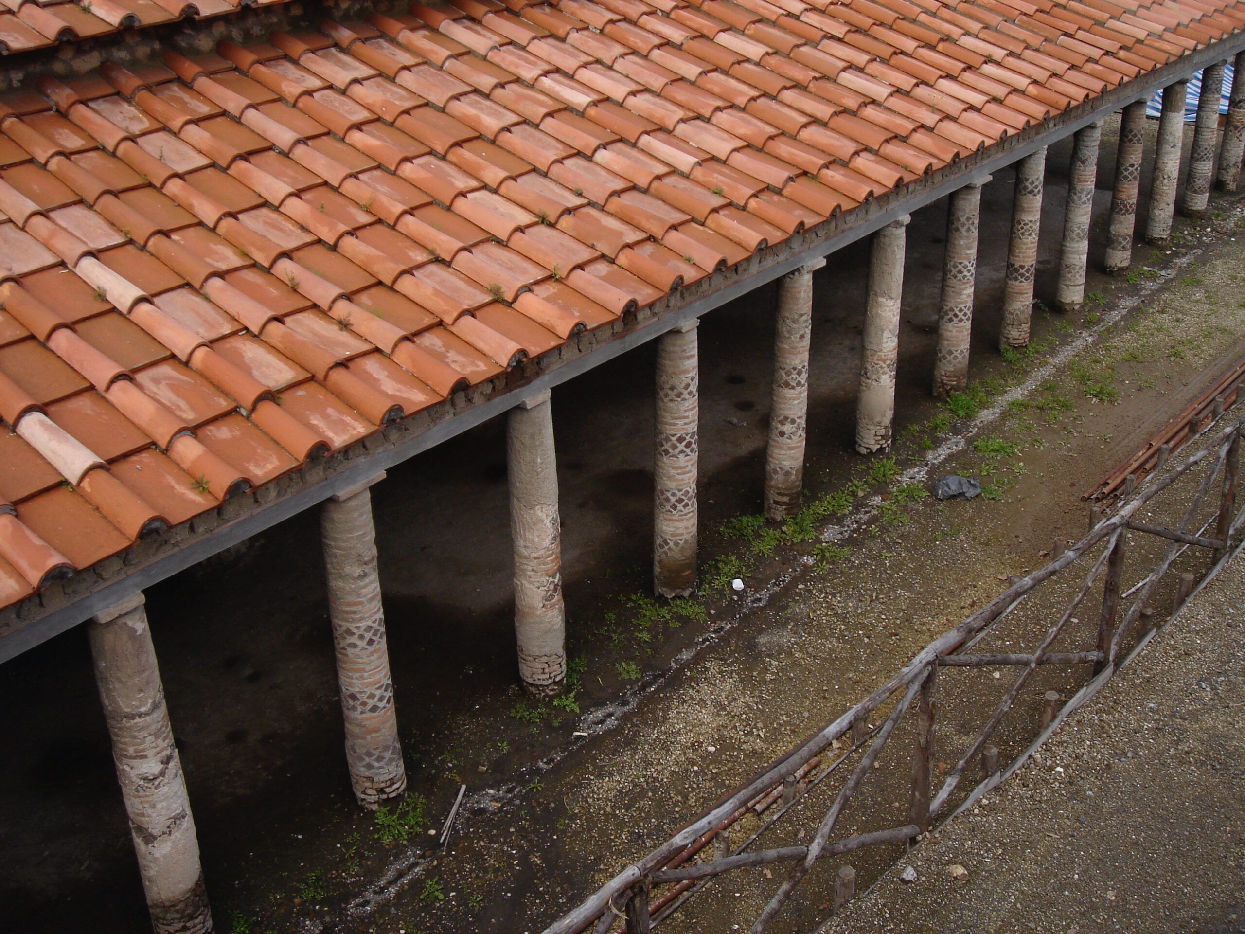Roofed portico supported by decorated stone columns in Pompeii, viewed from above with terracotta tiles and a dirt floor below.