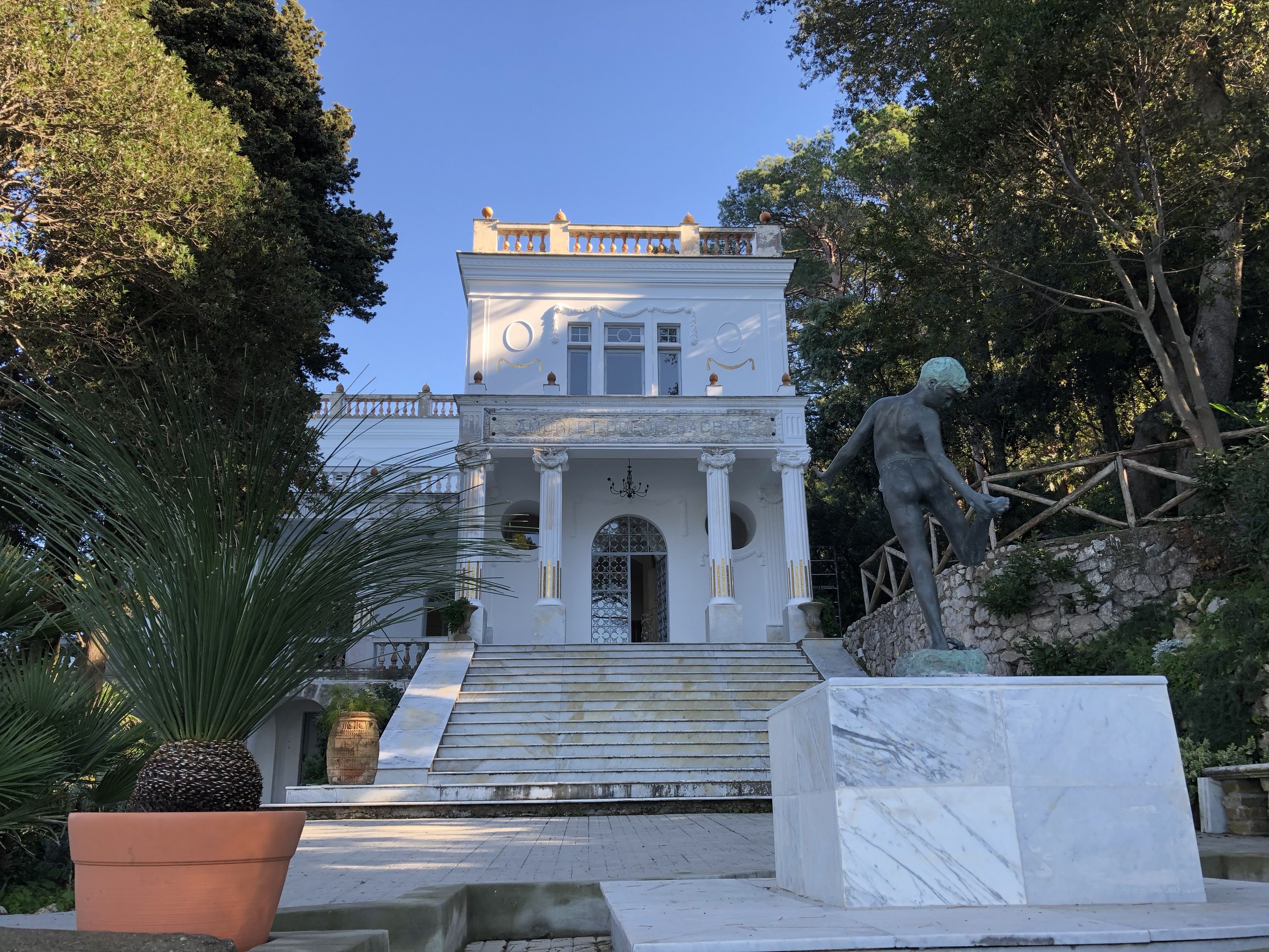 Grand entrance to Villa Lysis, a white neoclassical mansion with a central staircase and bronze statue in the garden.