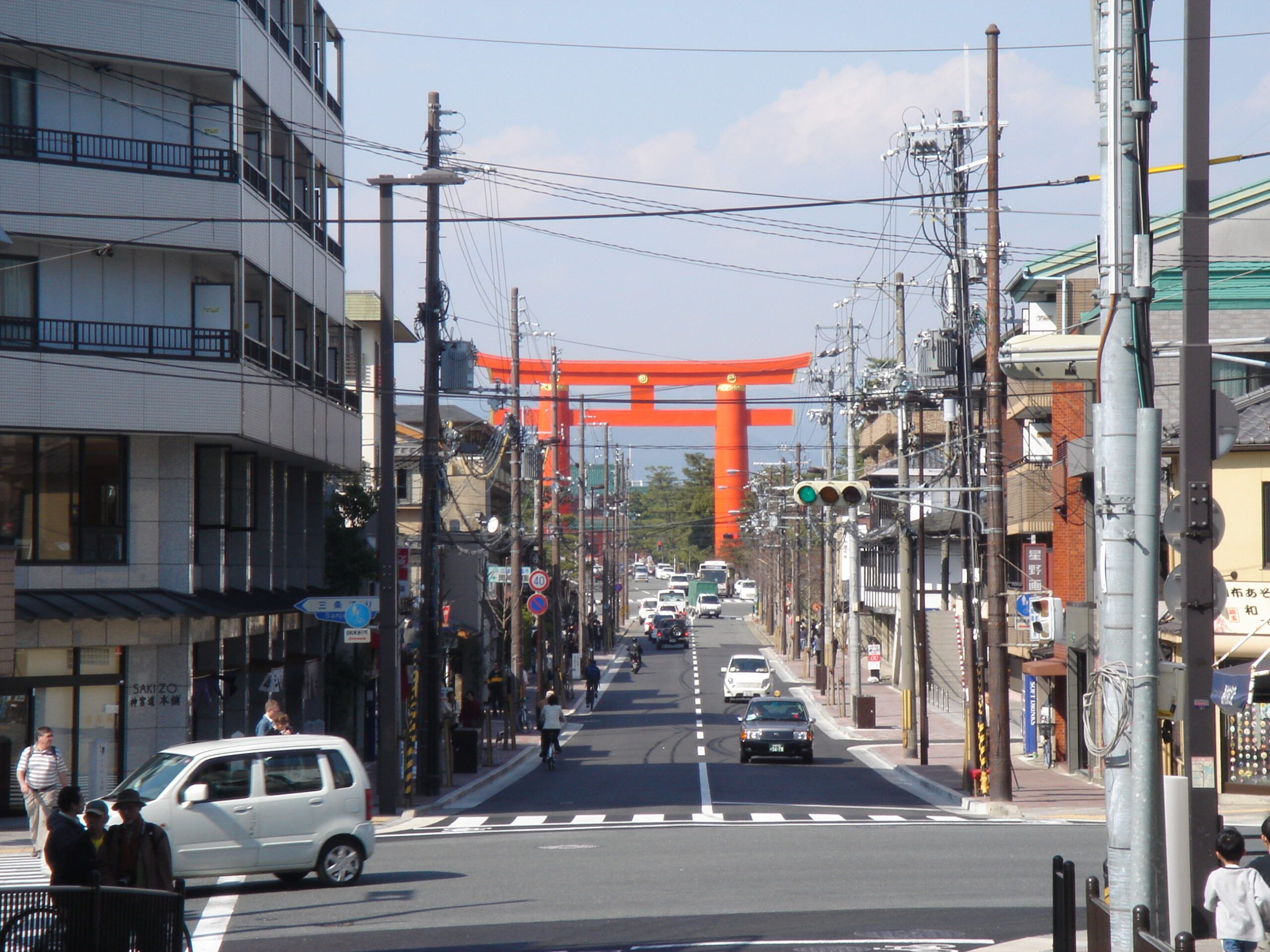 A busy street scene with traffic and power lines, leading toward two large red torii gates in the distance.