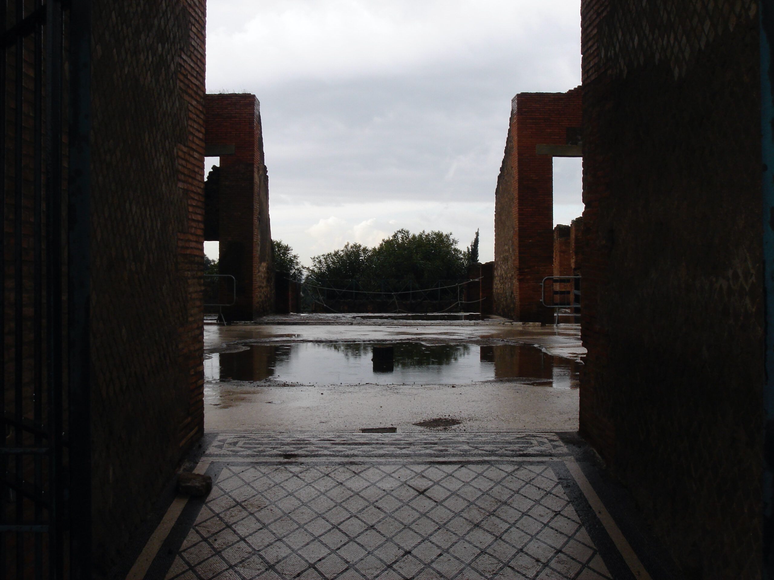 View through a doorway across a rain-slicked courtyard in Pompeii, with the geometric black-and-white tile floor still intact.