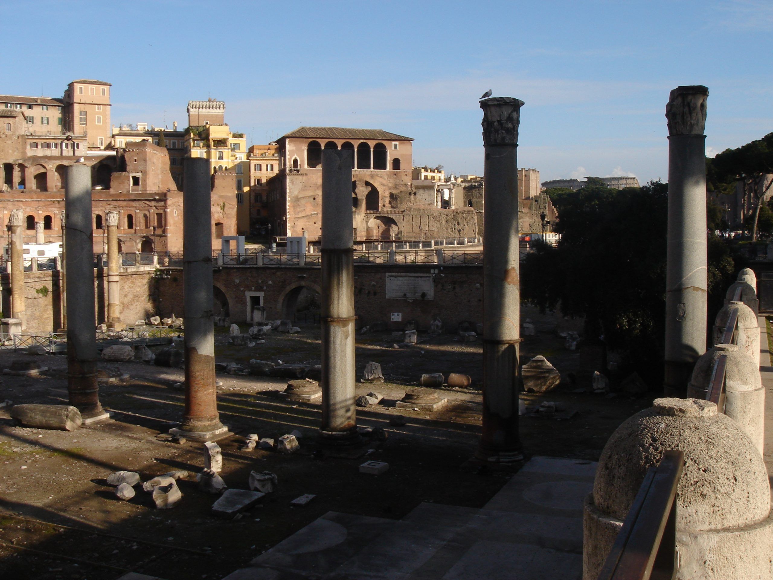 Another angle of the Forum of Trajan, showing broken columns and ruins with the markets of Trajan and city buildings beyond.