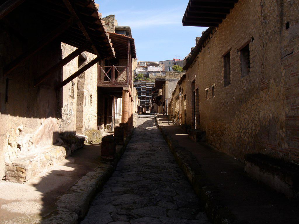 Narrow ancient Roman street in Herculaneum, lined with well-preserved walls, columns, and wooden balconies under reconstructed roofs.