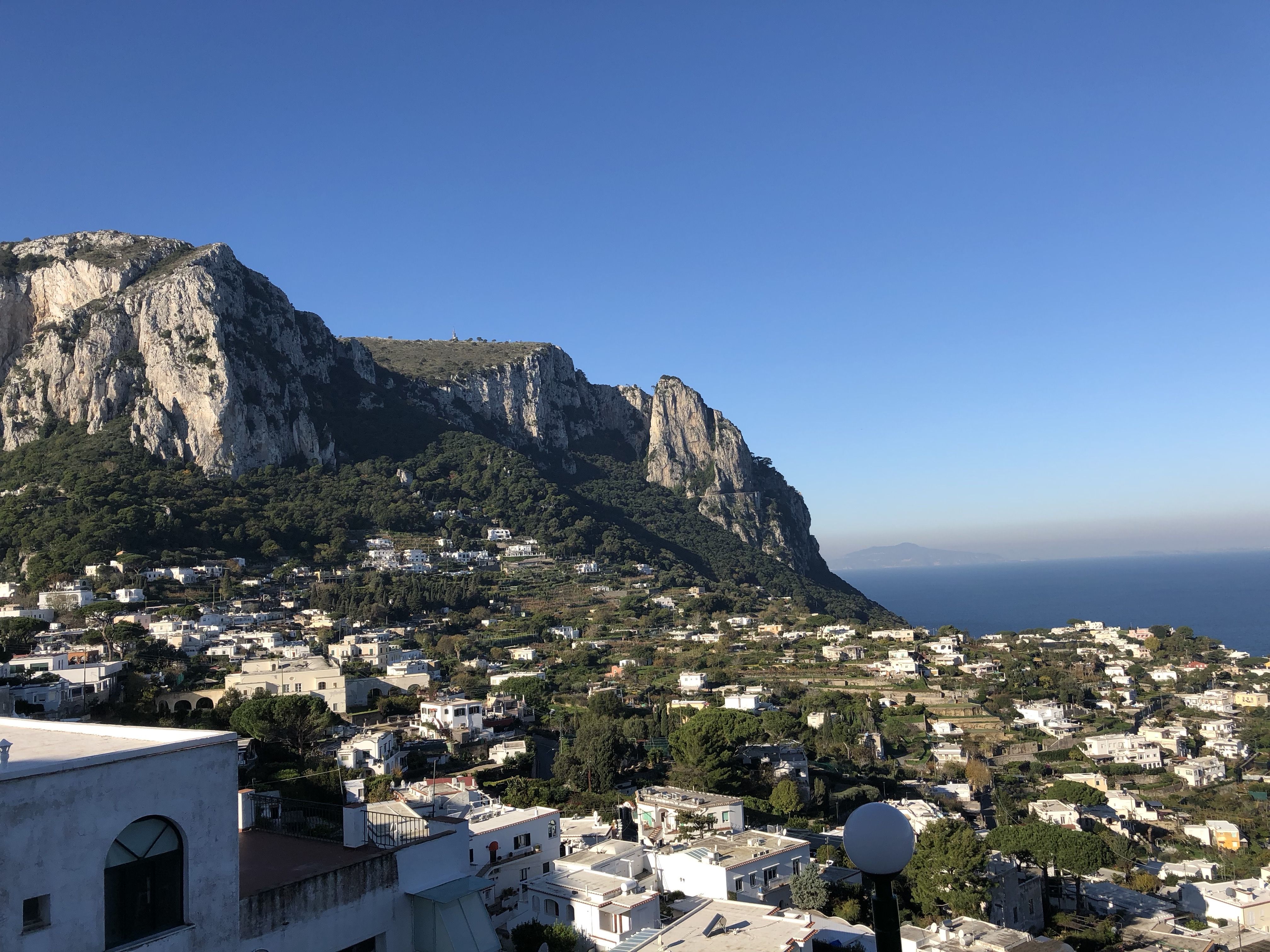 White houses and green hills spread below the rocky cliffs of Monte Solaro on Capri, under a clear blue sky.