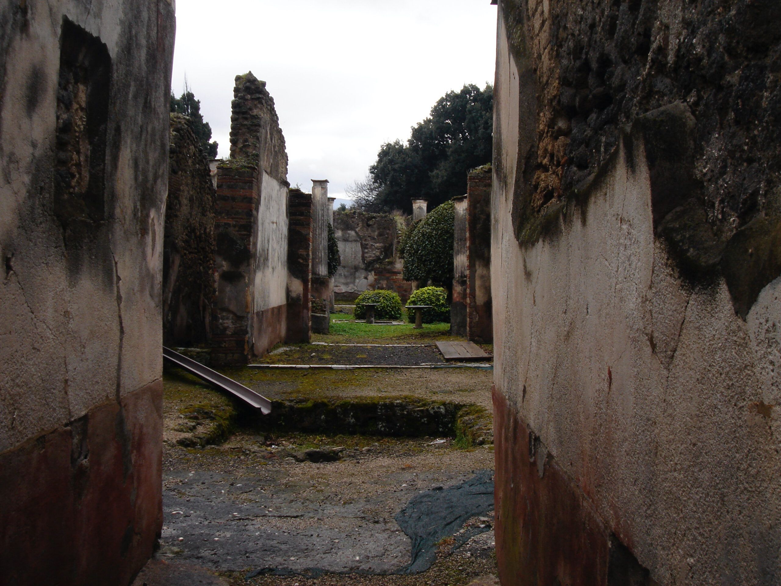 View into a ruined Roman house in Pompeii, with a garden courtyard visible beyond the weathered walls and scattered remains.