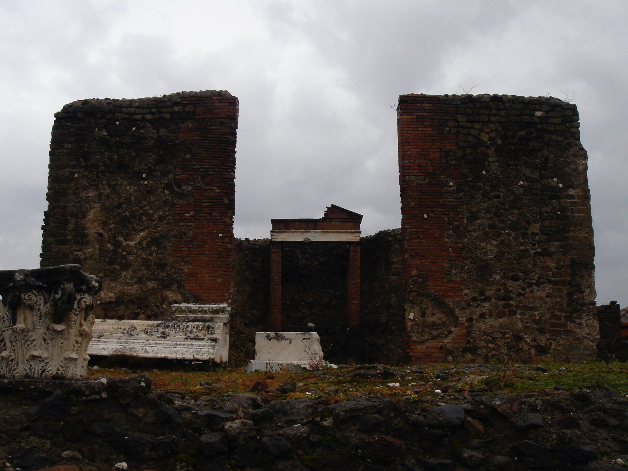 Two ruined brick towers and a central doorway mark the remains of a structure in the Macellum, with pieces of carved marble scattered nearby.