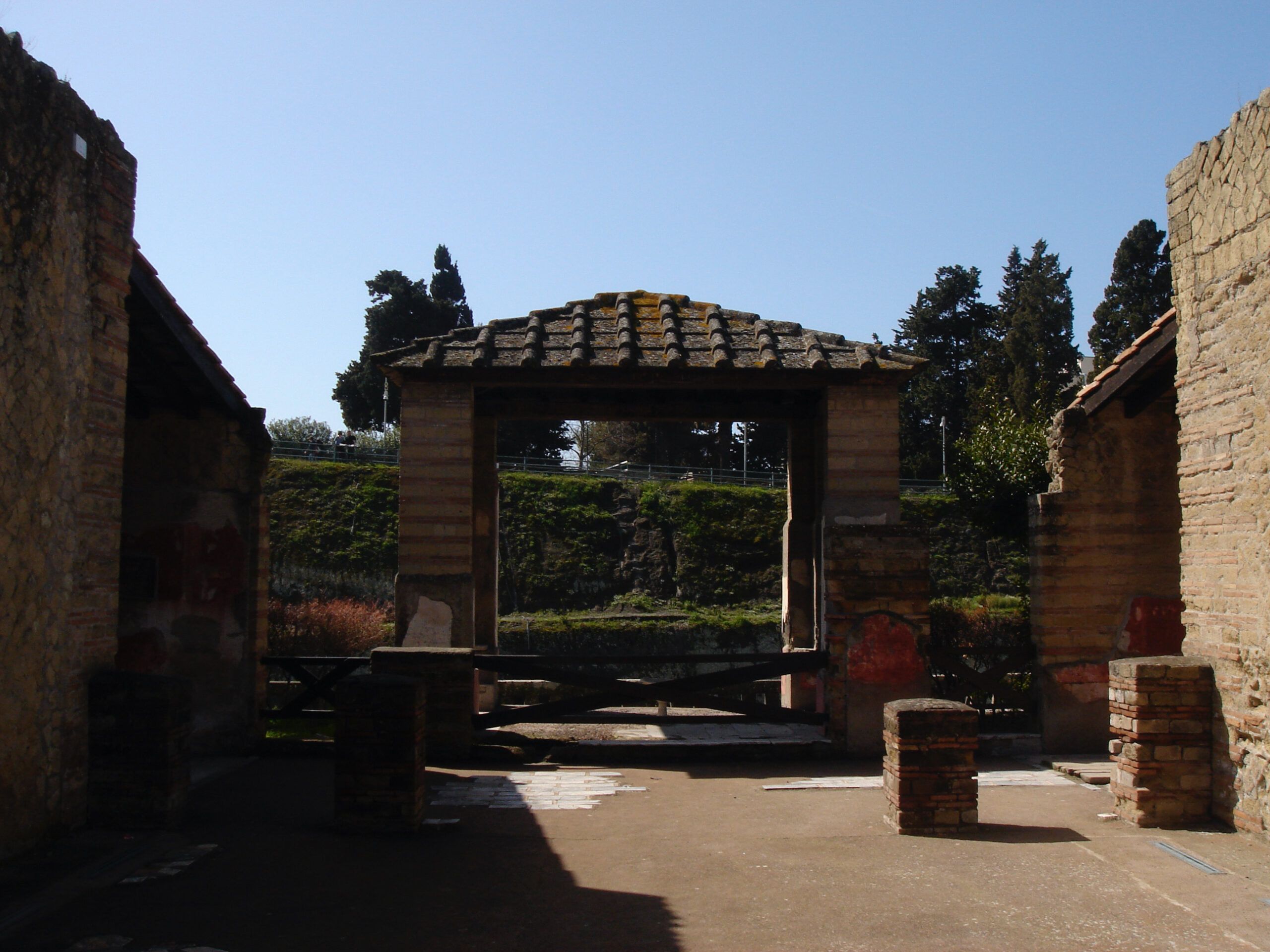 View of a roofed structure within a Roman house in Herculaneum, with the remains of painted walls and a tiled roof framed by brick columns.