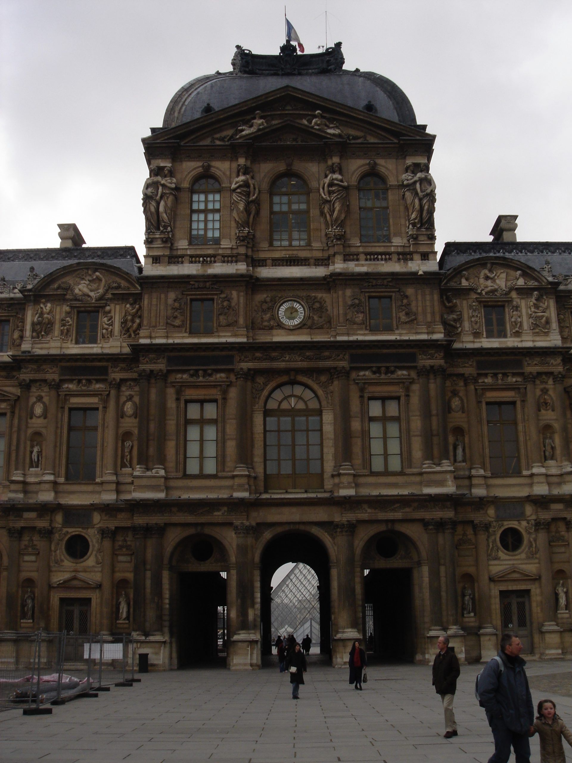 Ornate entrance of a Louvre courtyard building with sculptures and a view of the glass pyramid through the arch.