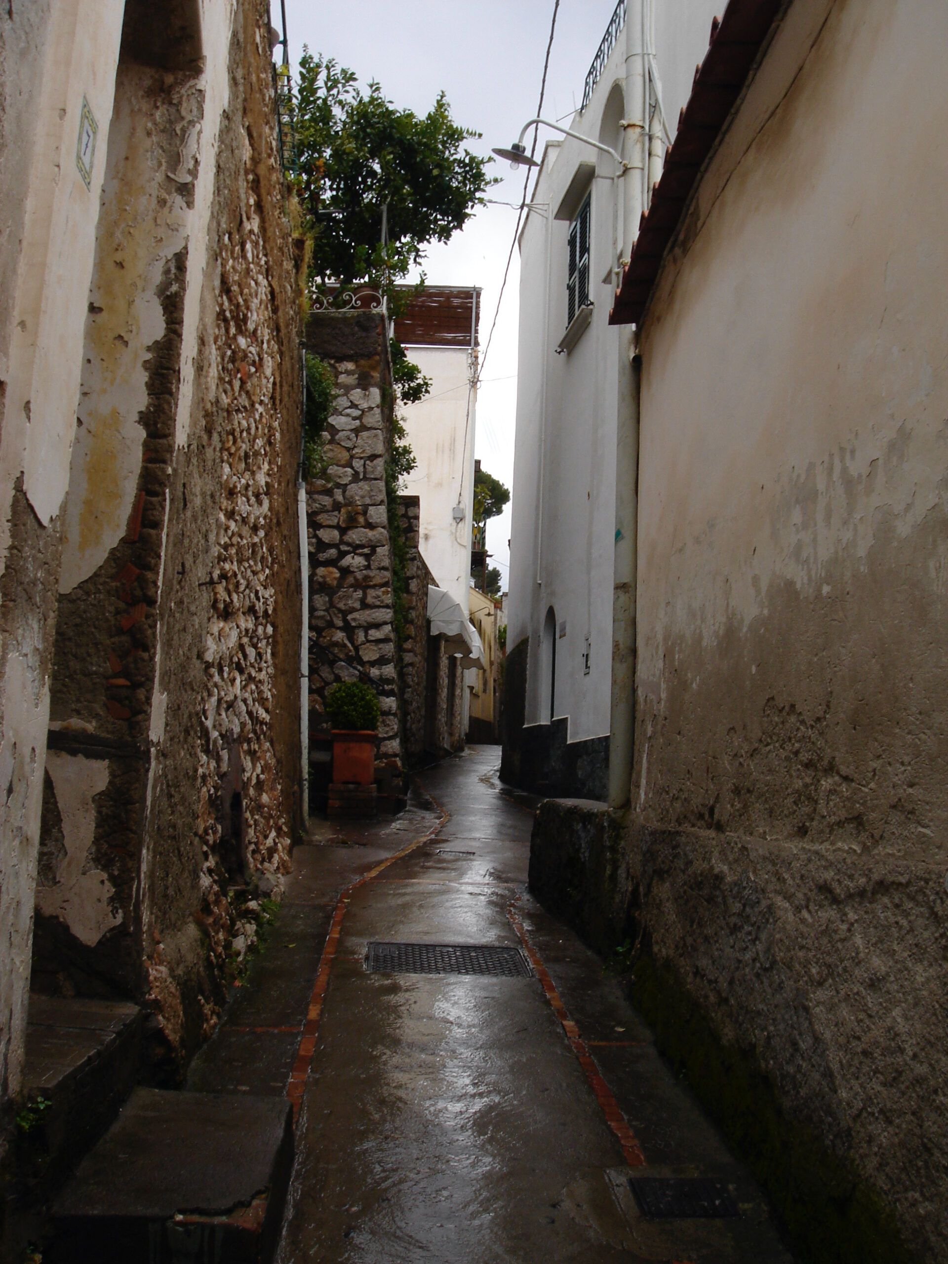 Narrow, rain-slicked alley between old stone and stucco buildings, leading uphill through a quiet village.