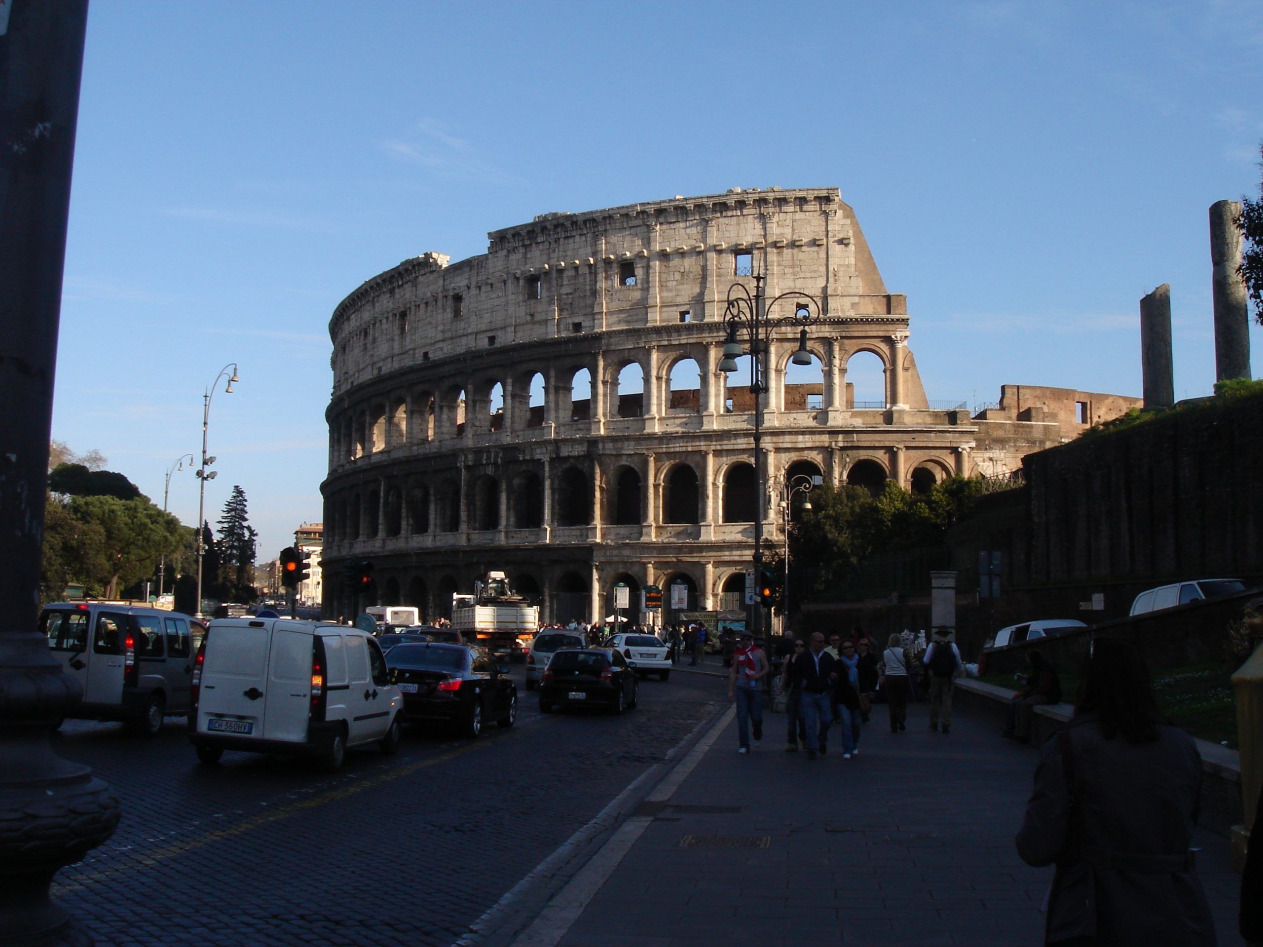 The Colosseum in Rome, viewed from the street with traffic and pedestrians in the foreground on a sunny afternoon.