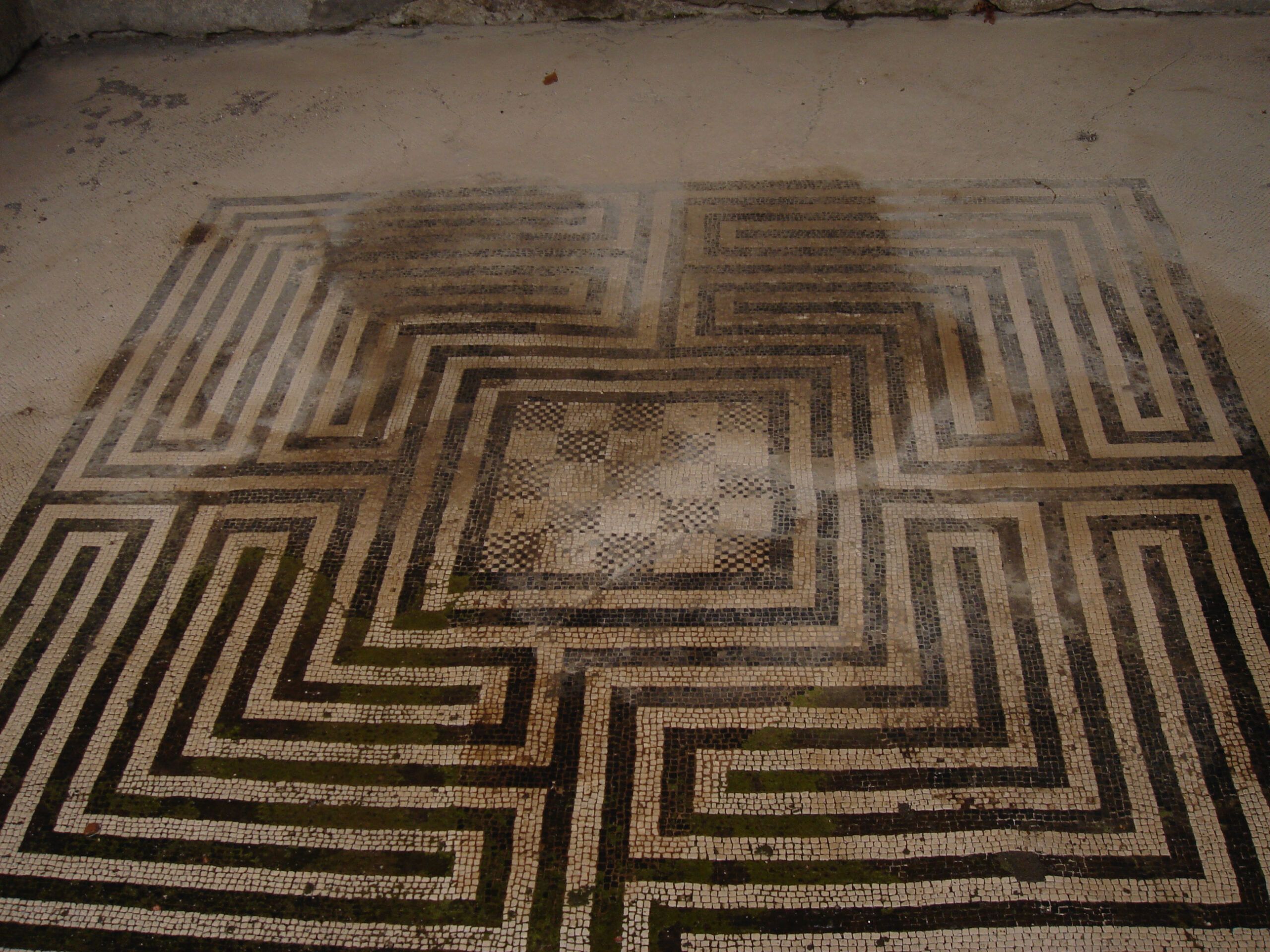 Intricate black-and-white mosaic floor in Pompeii, with a maze-like square pattern partially darkened by water and age.