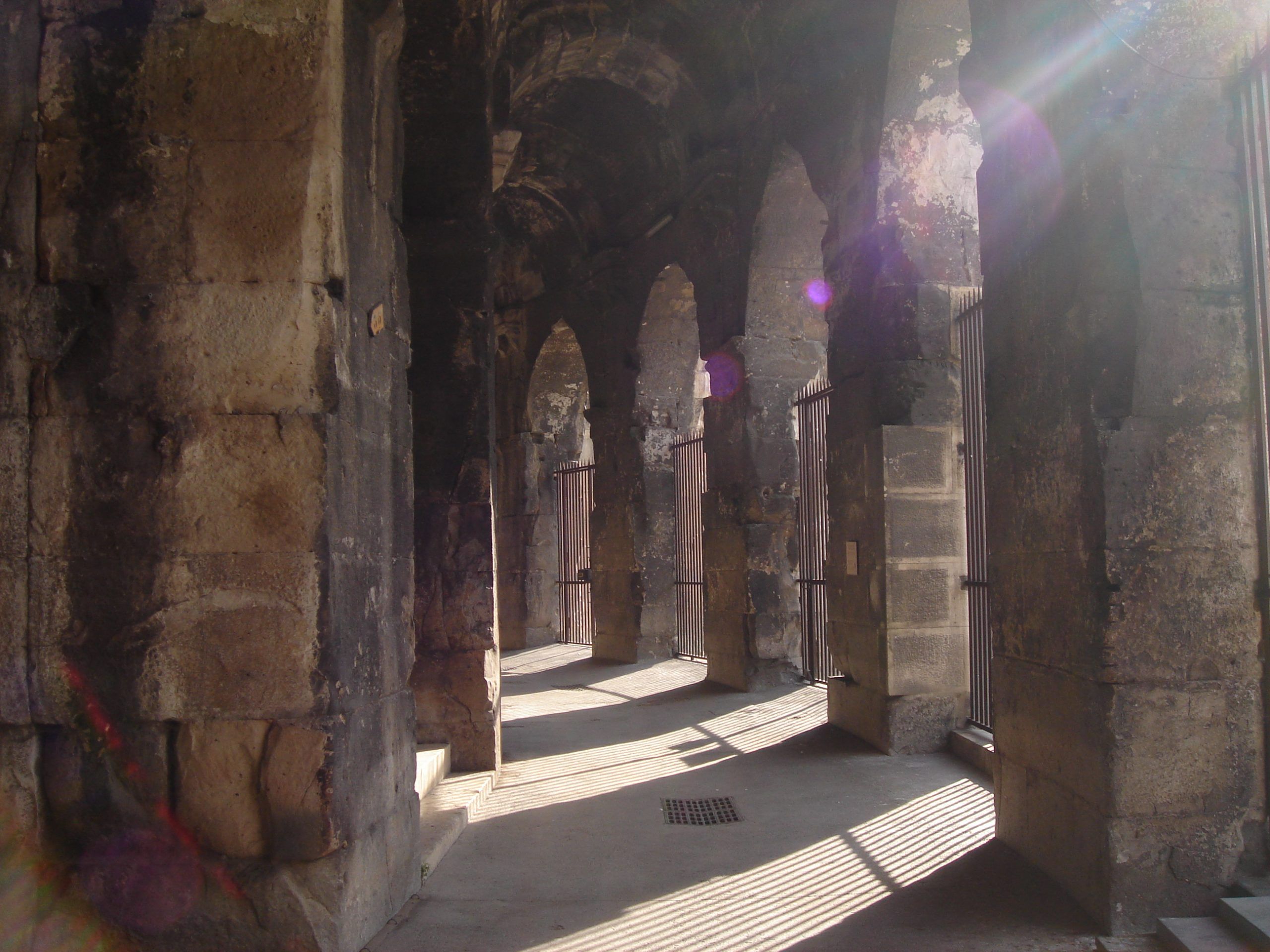 Sunlit corridor inside the Roman amphitheatre in Nîmes, with shadows cast through the arches and barred gates.