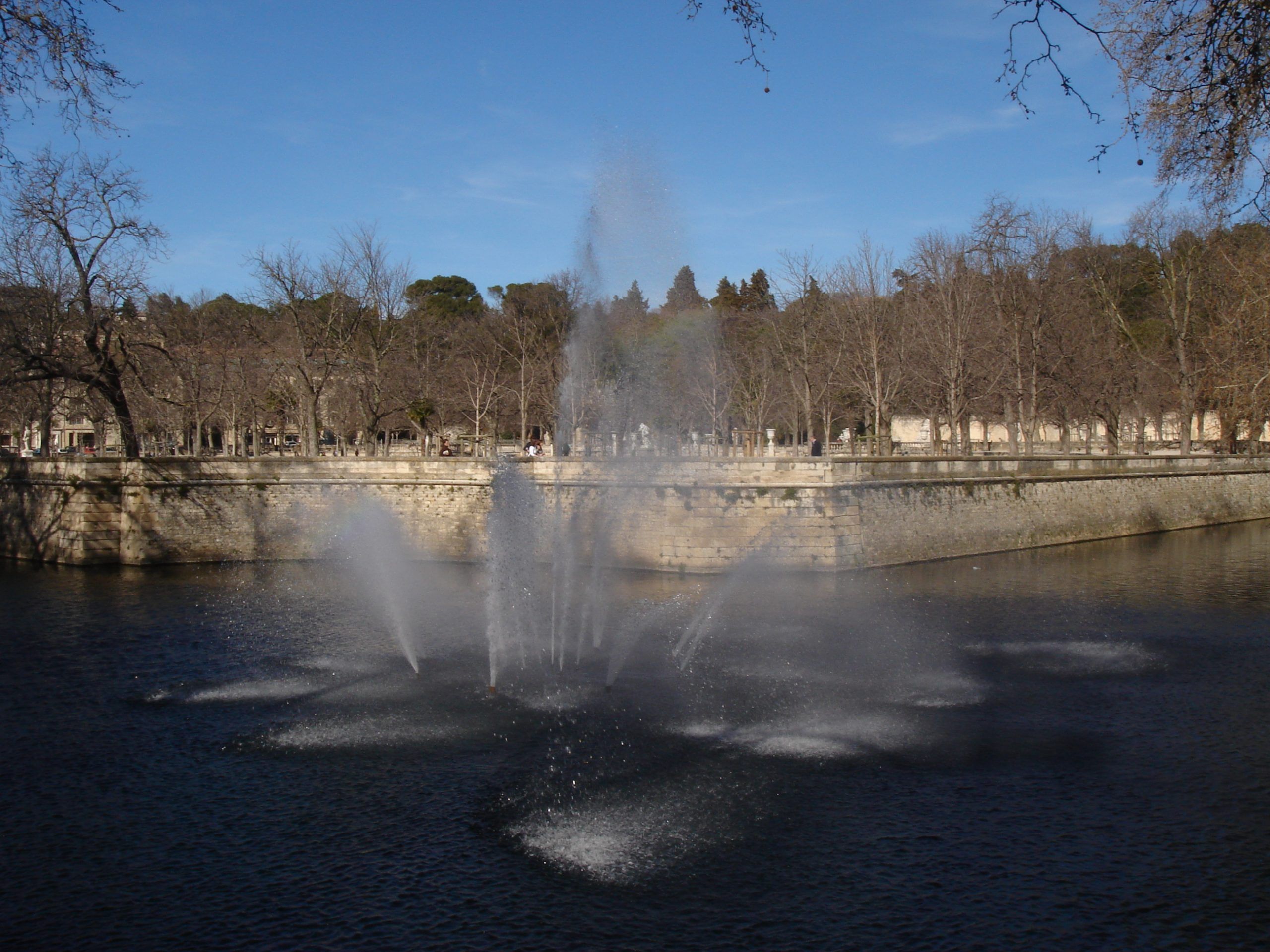 Water fountain spraying in the centre of a park pond surrounded by leafless trees in Nîmes.