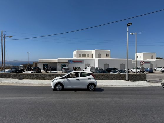 Across the road is another low white building with a flat roof and rounded corners. It's a big shop. The sign on it says AB, and there's some other Greek writing underneath. In front of the shop there's a car park with a dozen cars in it.