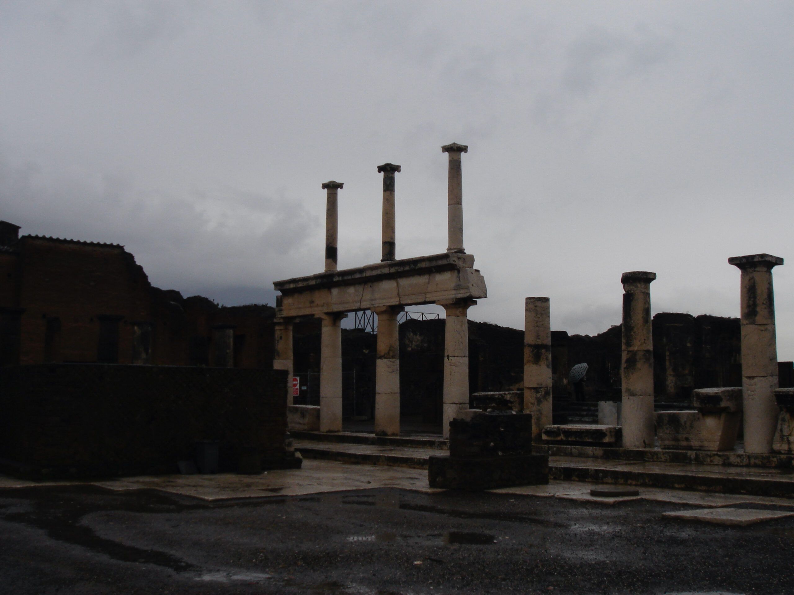 Columns of the Temple of Vespasian in Pompeii stand against a gloomy sky, their marble surfaces weathered by time.