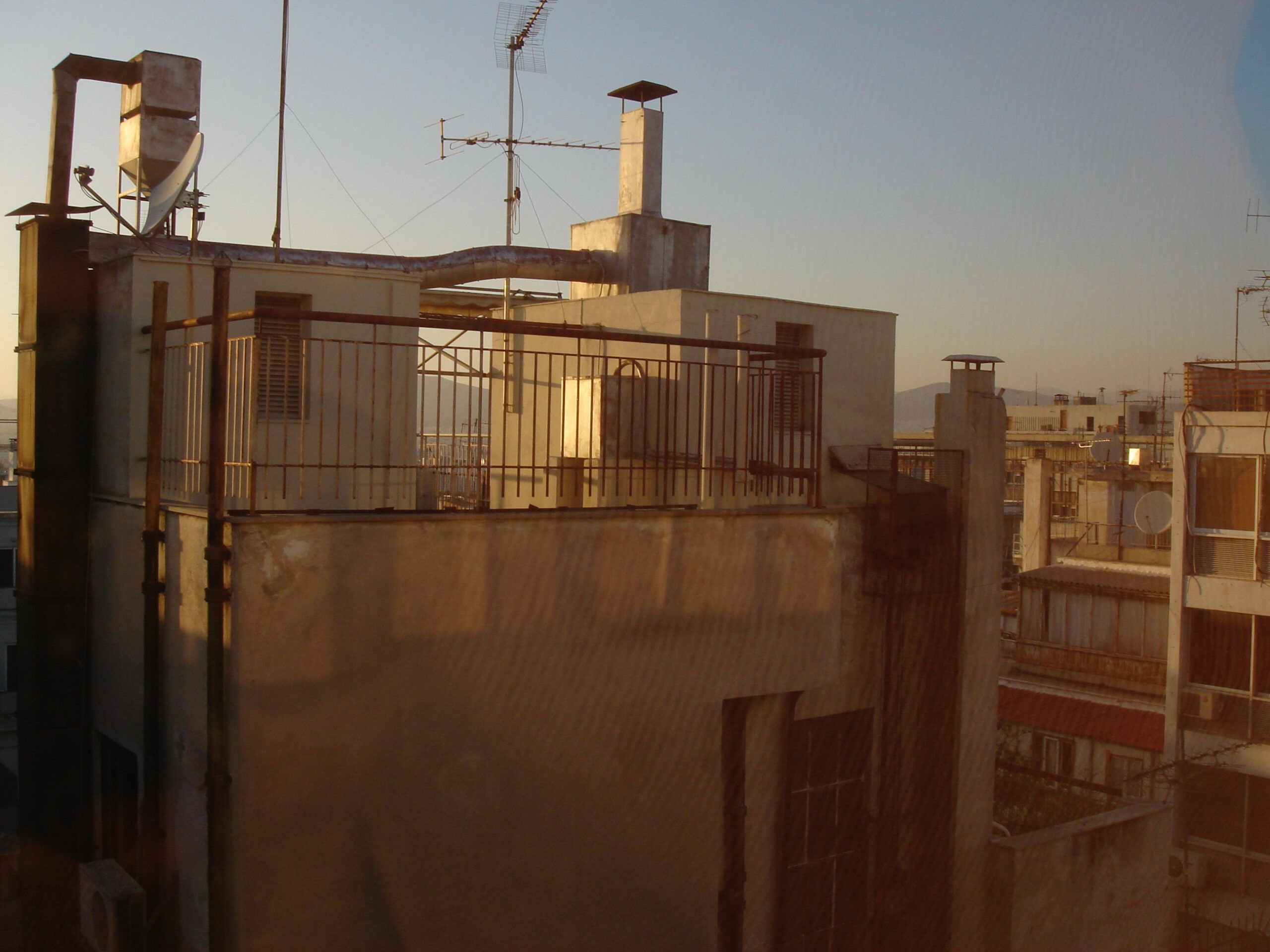 Rooftop view of an apartment building in Athens at golden hour, with satellite dishes and antennas silhouetted against the sky.