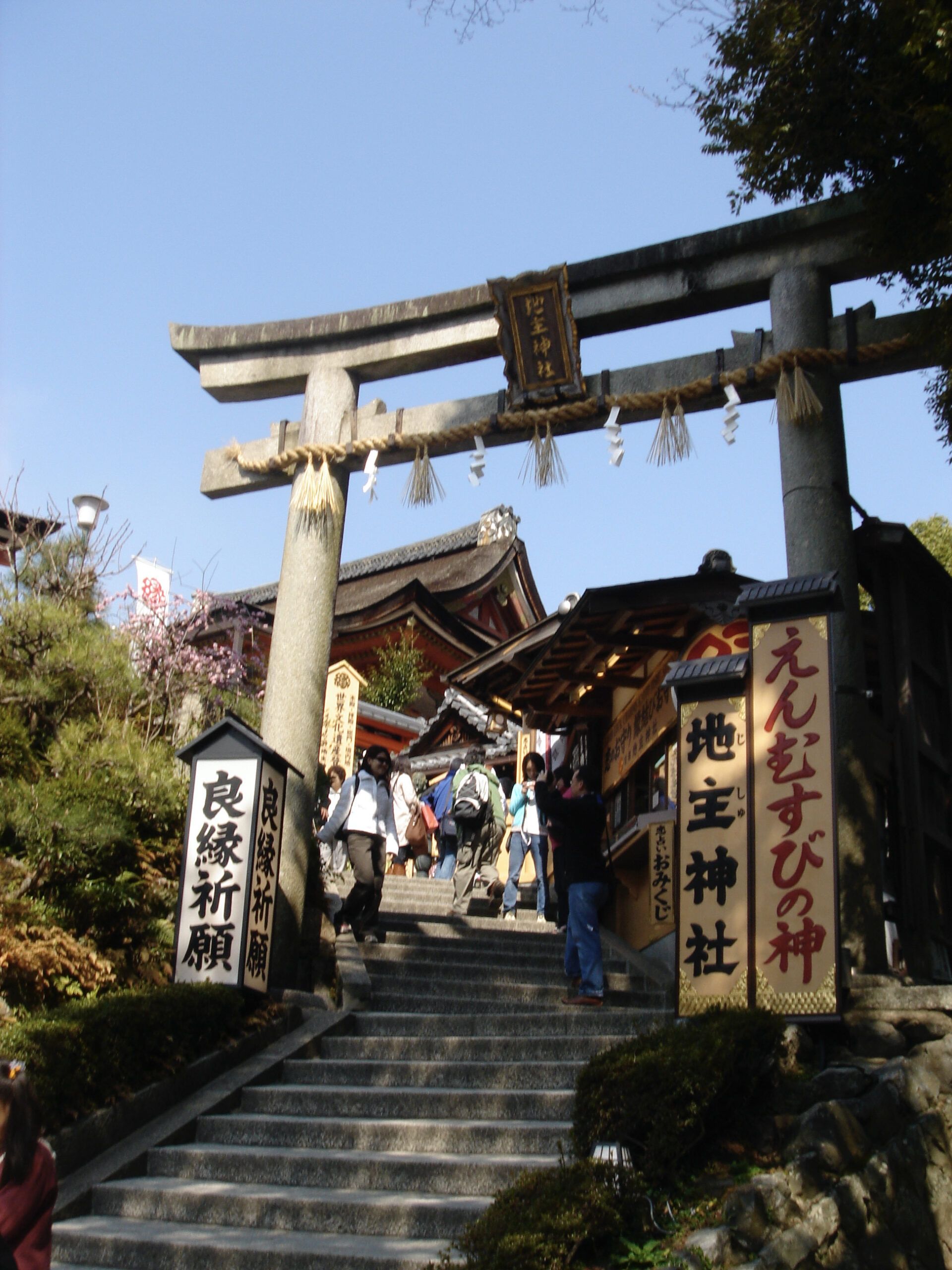 A large stone torii gate at the entrance to a shrine, with visitors ascending the steps past signs and hanging ropes.