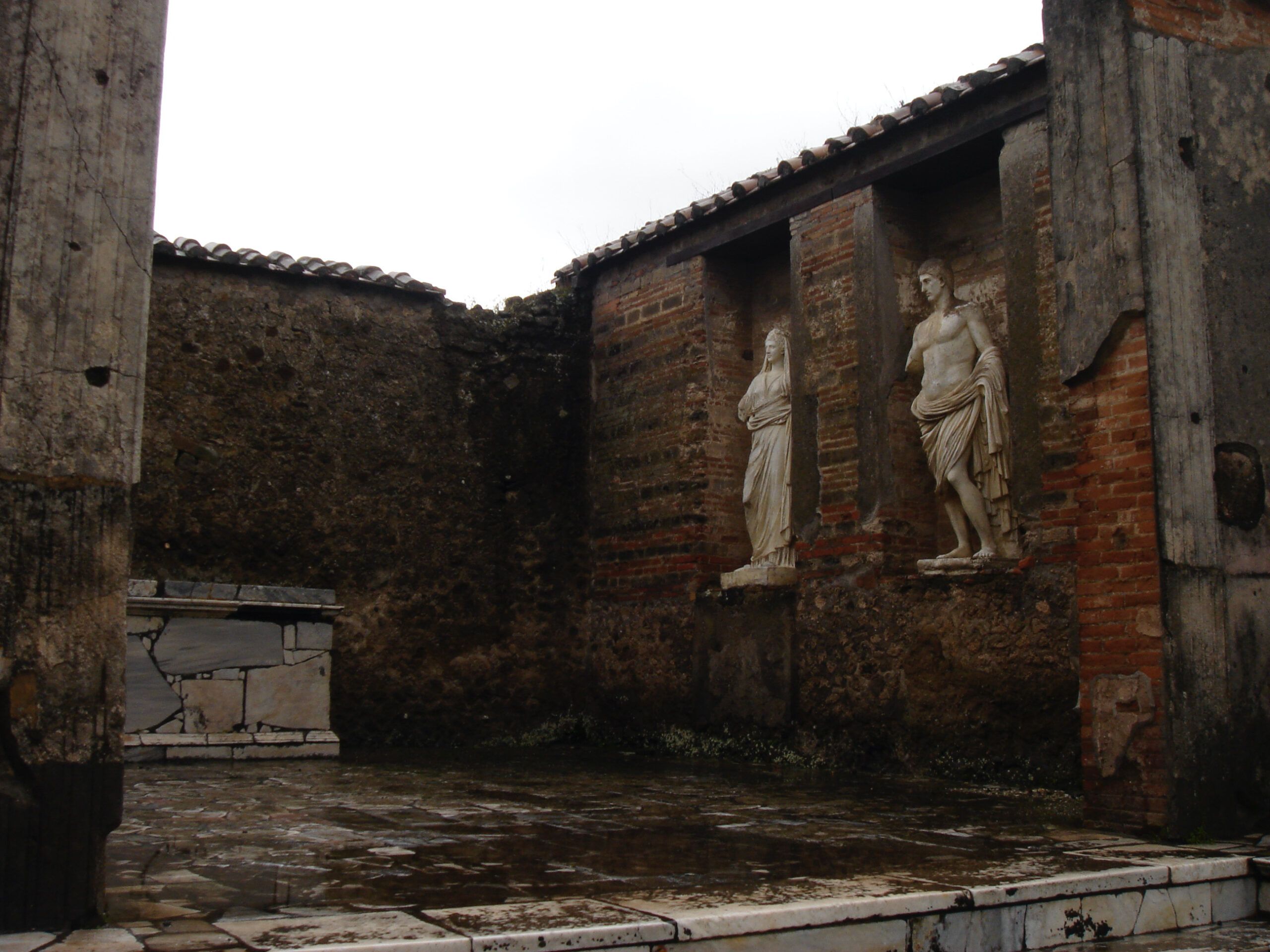 Two marble statues in niches decorate the wall of a Roman house in Pompeii, surrounded by dark brick and columns.
