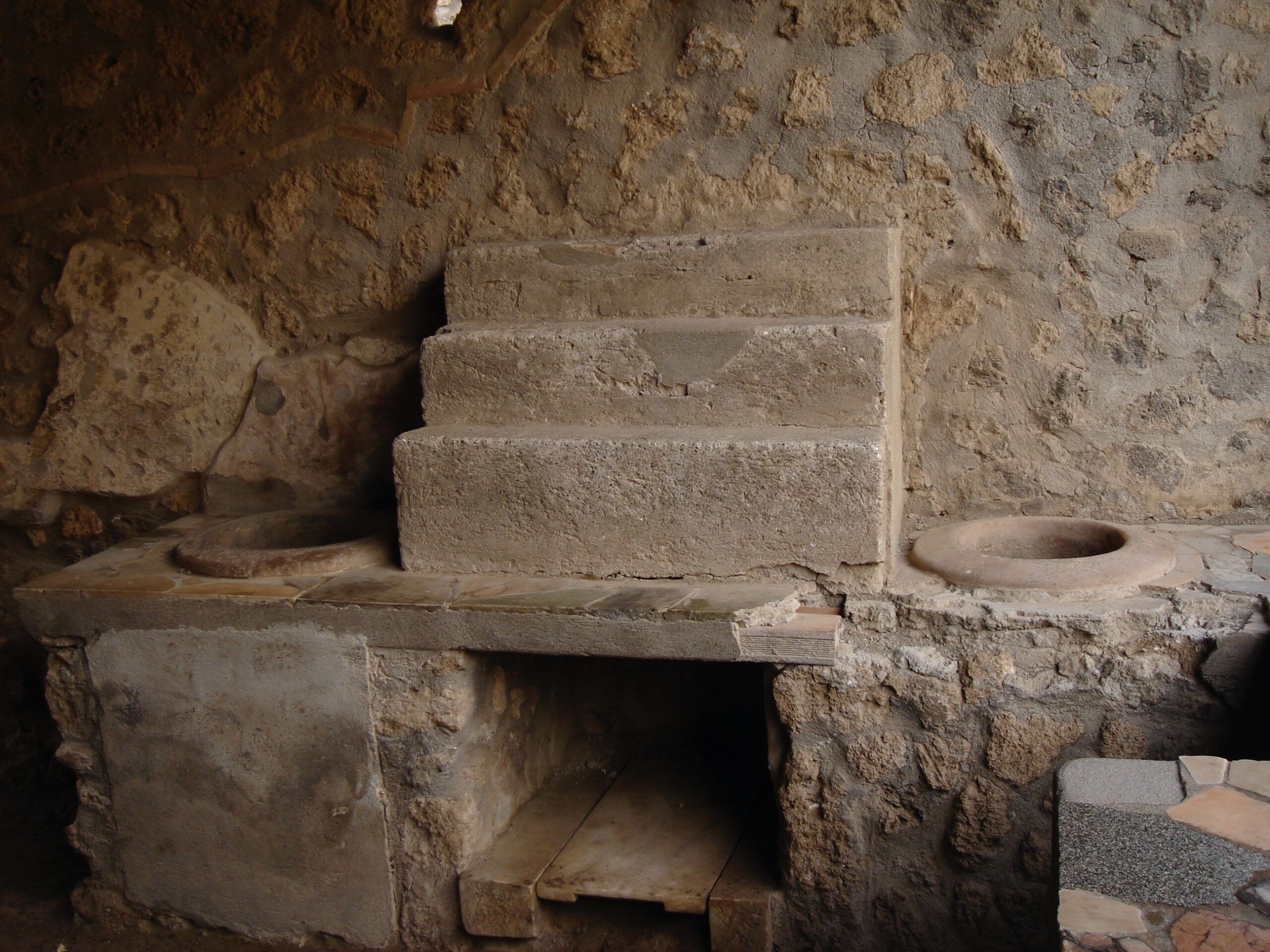 Stone counter with built-in dolia from a Pompeian thermopolium, featuring worn surfaces and masonry embedded in plaster walls.