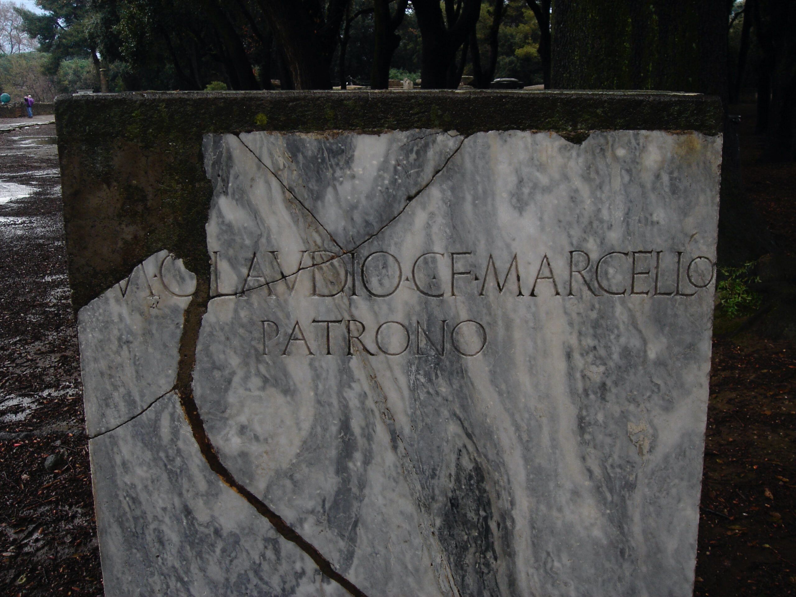 Broken marble inscription stone reads “M. CLAUDIO C. F. MARCELLO PATRONO” standing at the edge of the Triangular Forum in Pompeii.