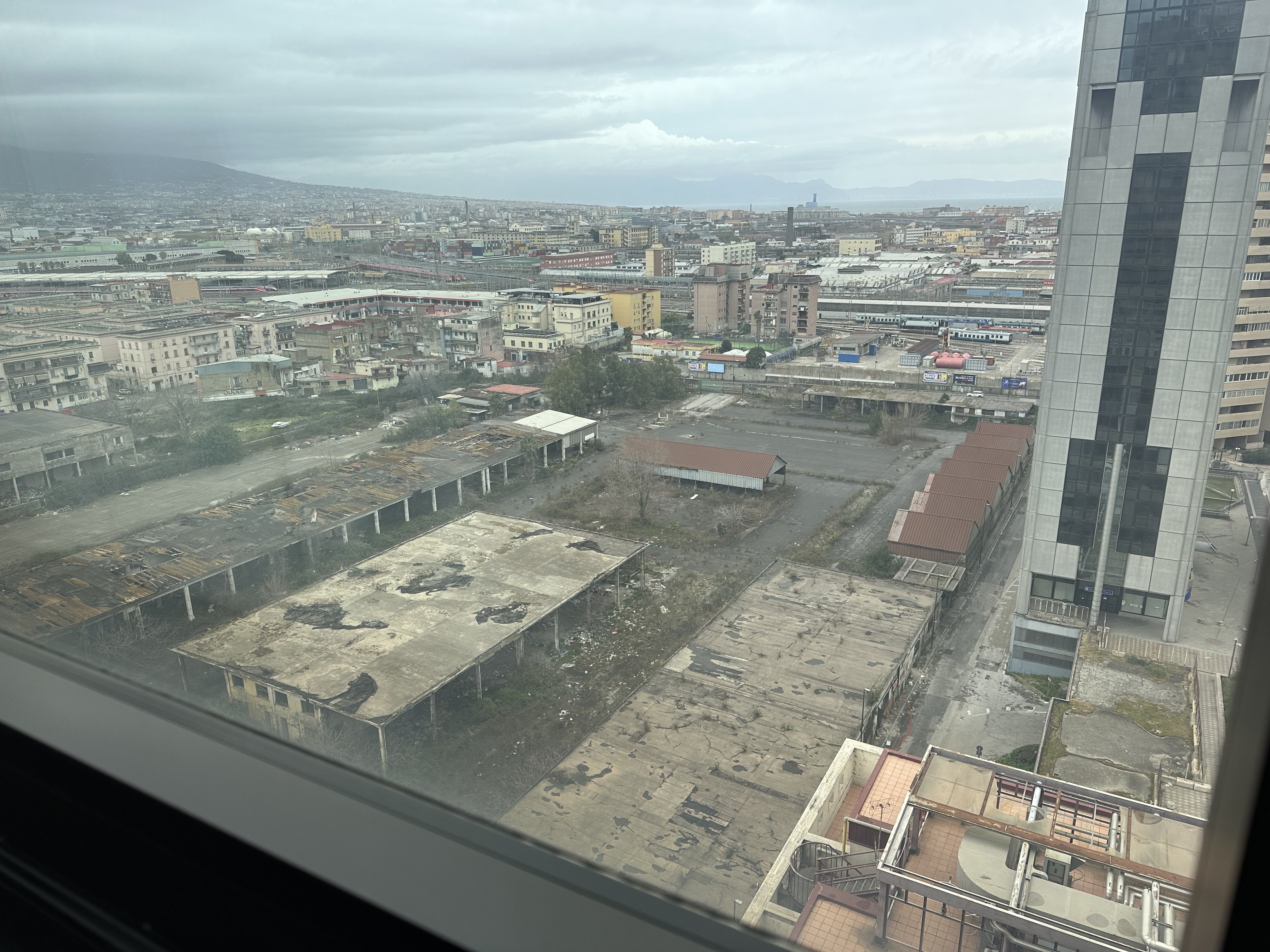 View from a high-rise hotel window overlooking industrial buildings, apartment blocks, and Mount Vesuvius in the distance.
