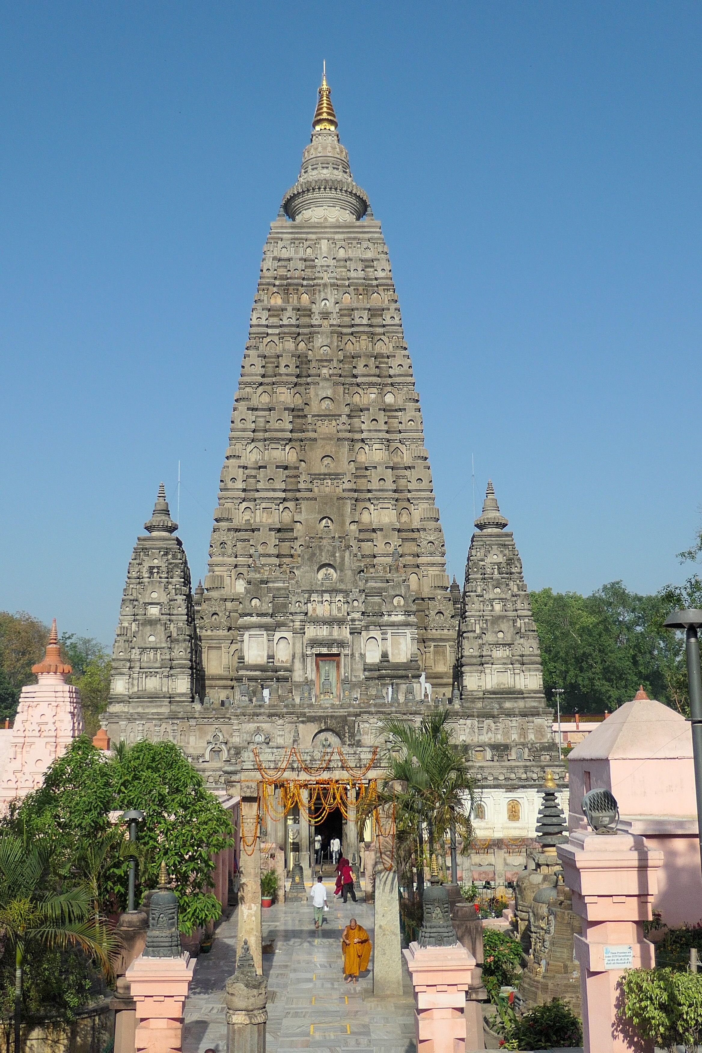 View of the Mahabodhi Temple in Bodhgaya, a tall pyramidal structure with ornate carvings and a golden spire, under a clear blue sky.