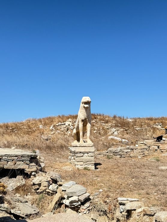 It's a dry scrubby landscape under a bright Mediterranean sky. On a stone plinth is a weathered statue of a lion, its mouth open.