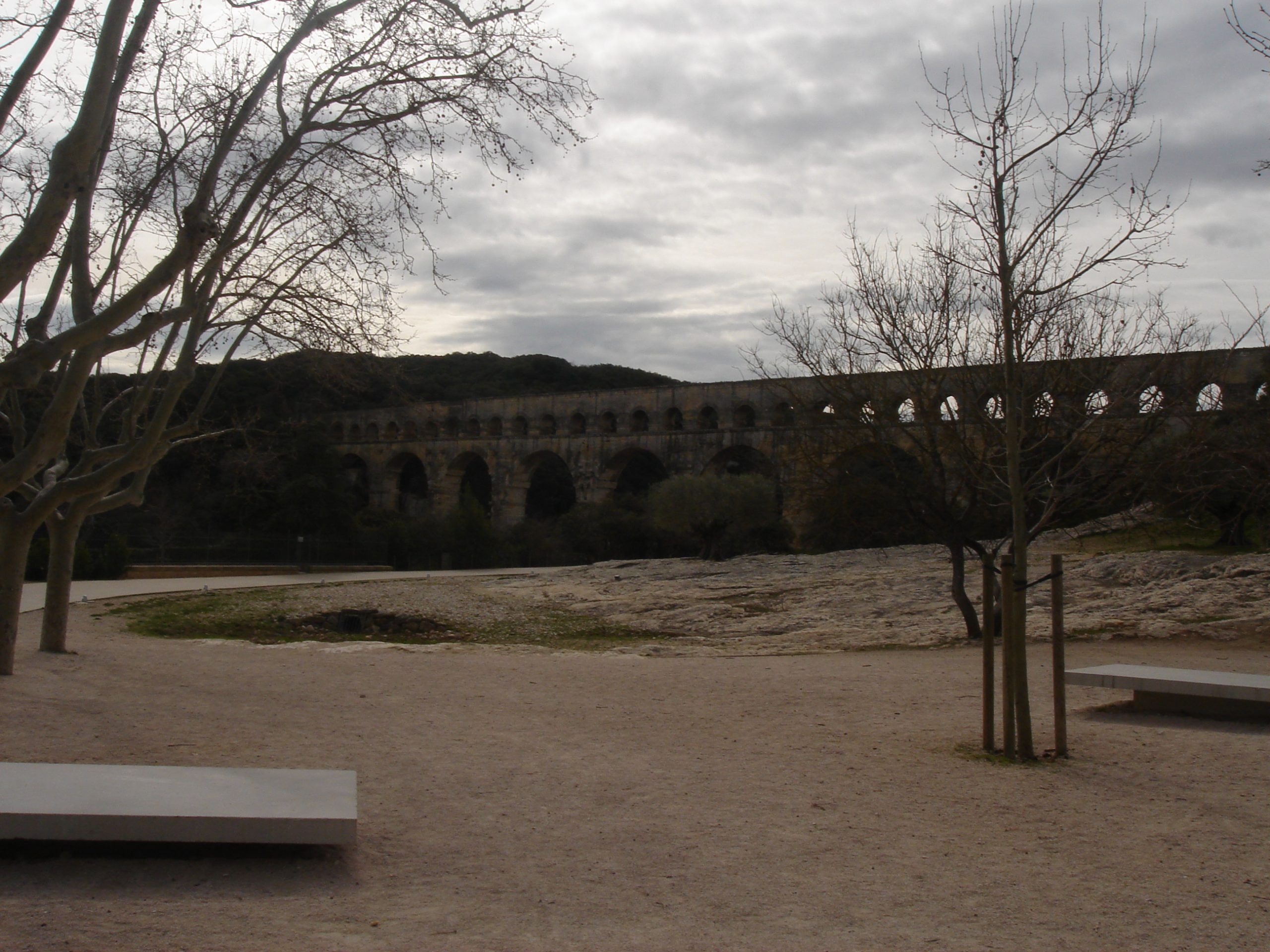Distant view of the Pont du Gard aqueduct stretching across a wooded valley, seen from a rocky foreground with bare trees and benches.