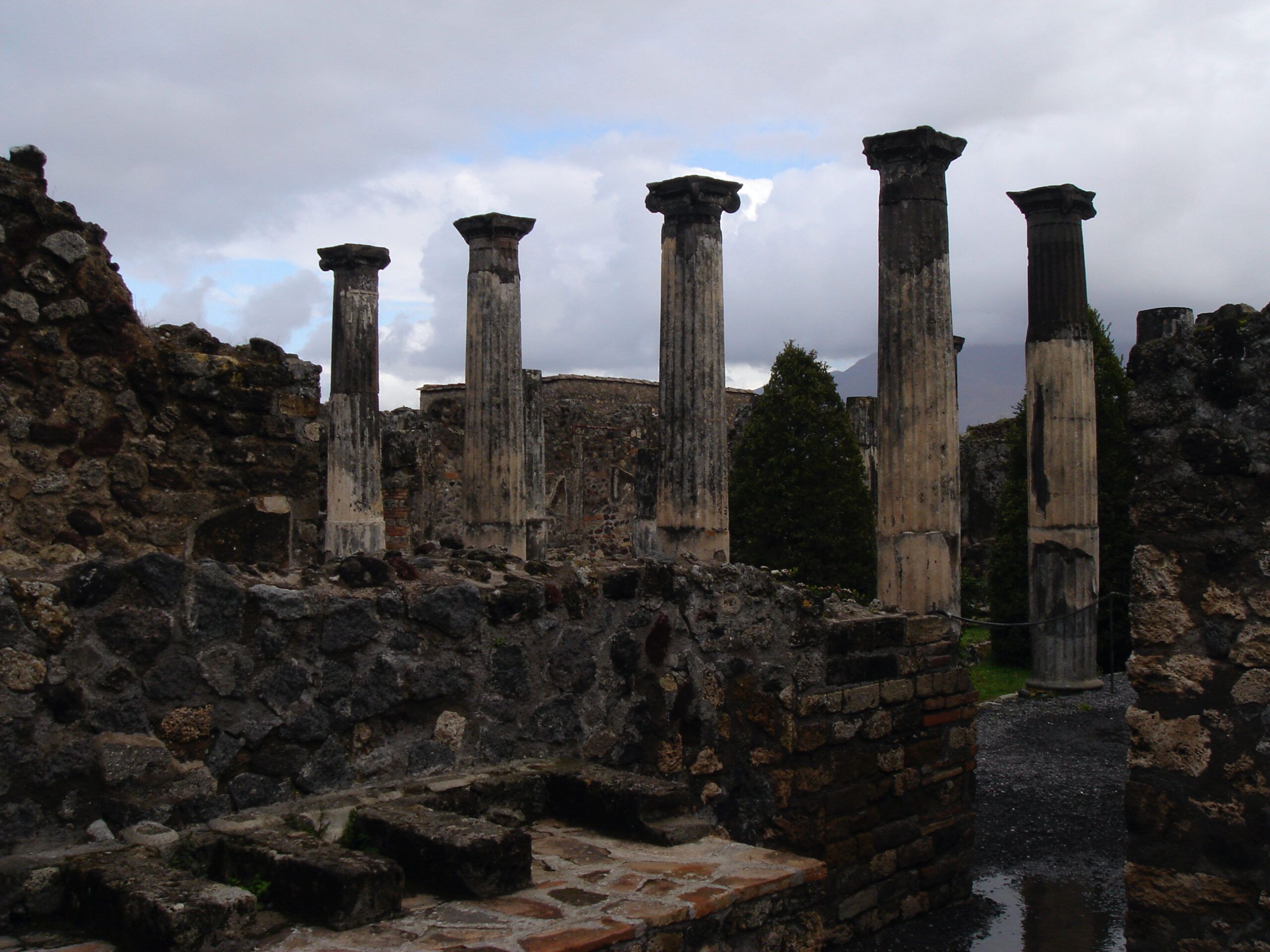 View of standing columns in the Forum of Pompeii with dark stone ruins in the foreground and Mount Vesuvius faintly visible in the distance.