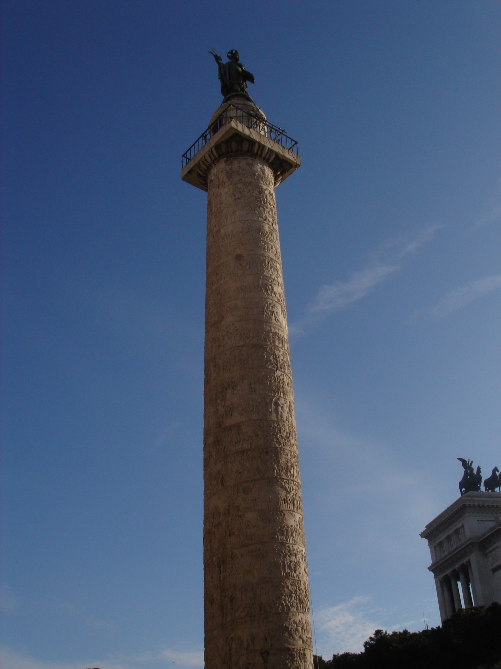 A distant view of Trajan’s Column rising against a clear blue sky, topped with a statue and decorated with a spiral relief.