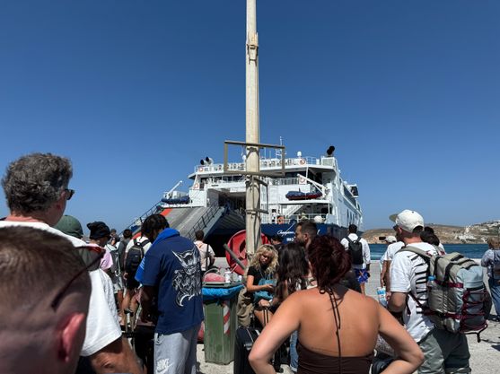 A big crowd of people standing in the blazing sun, waiting for the huge ferry that has just appeared behind them.
