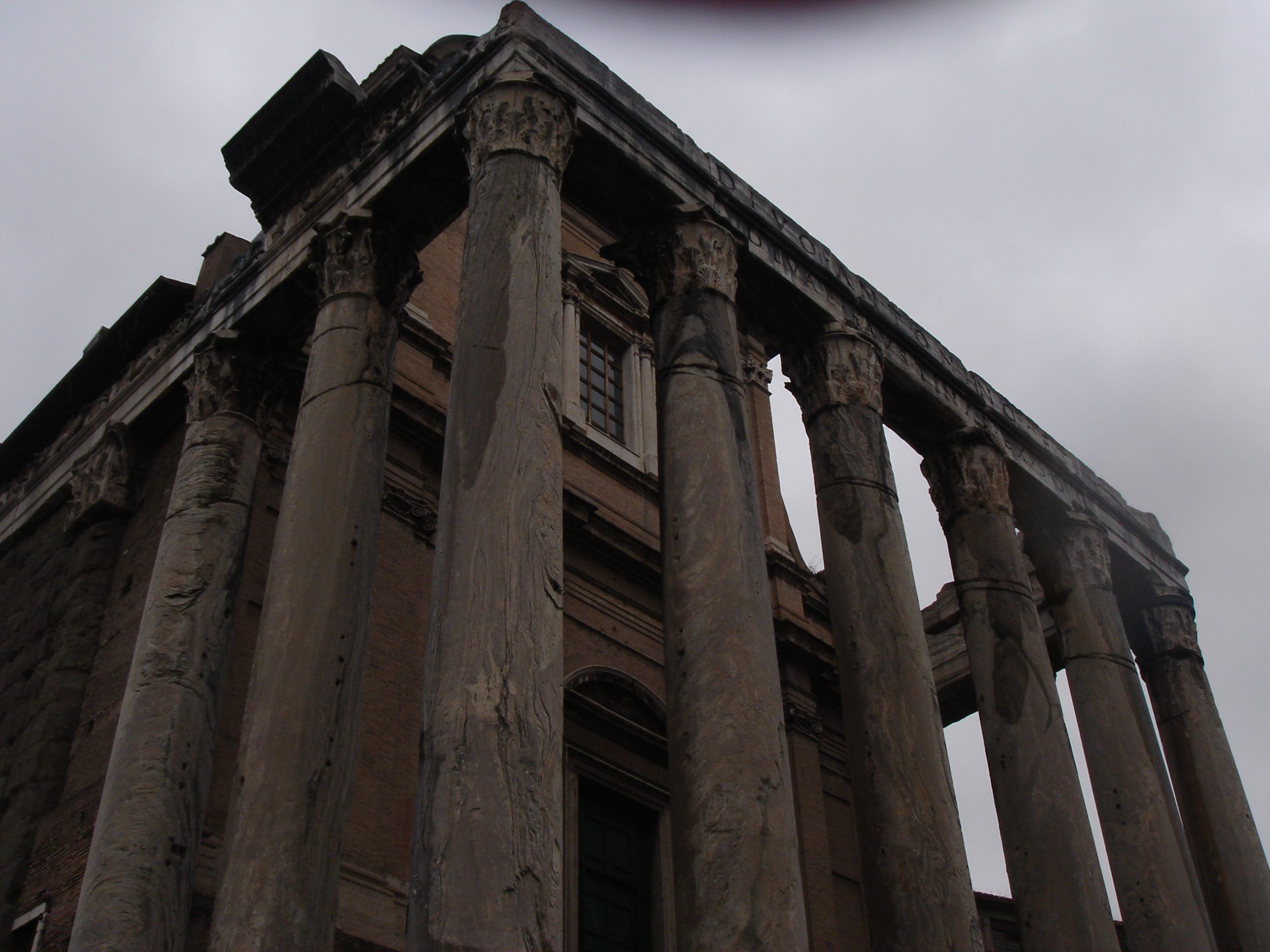Angled view of the Temple of Antoninus and Faustina in the Roman Forum, with worn marble columns supporting an entablature inscribed in Latin.