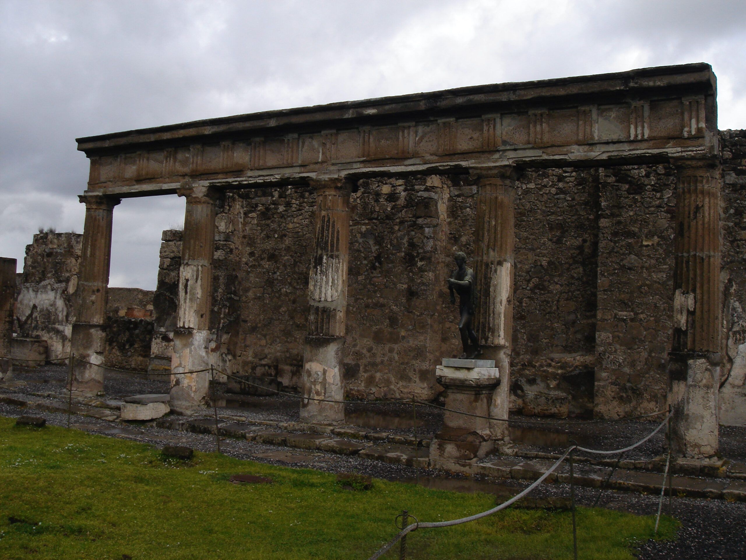 Statue of Apollo amid the ruins of his temple in Pompeii, with a colonnade and cloudy sky in the background.