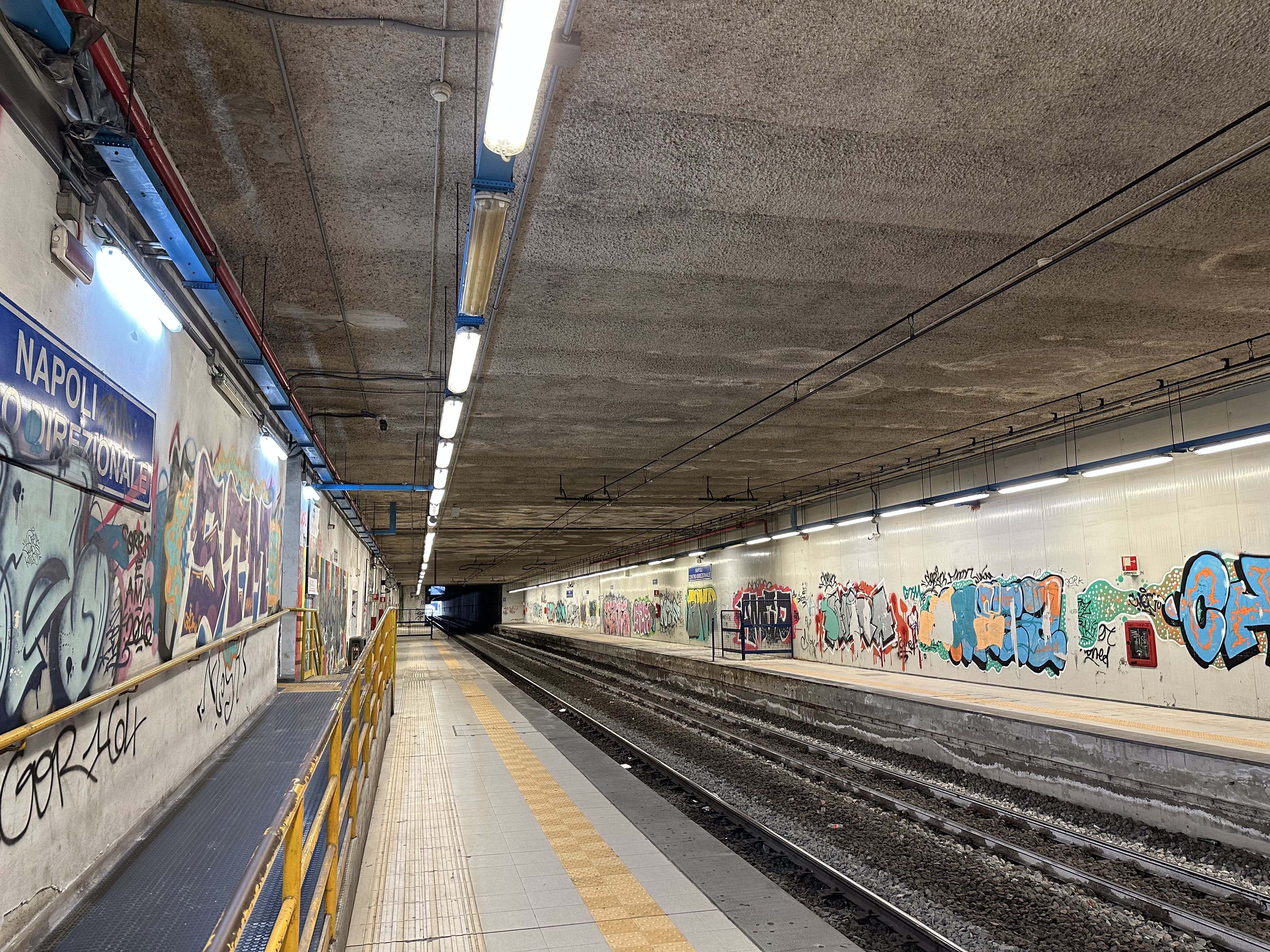Graffiti-covered underground train station in Naples, with empty tracks and no trains or people on the platform.
