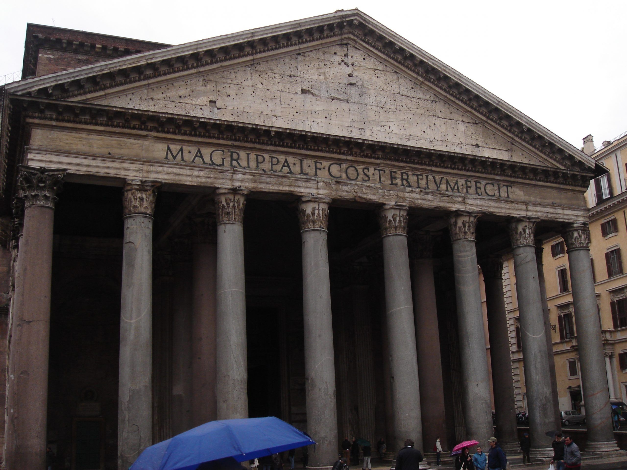 Front façade of the Pantheon in Rome with its iconic inscription and Corinthian columns, as visitors walk past with umbrellas.
